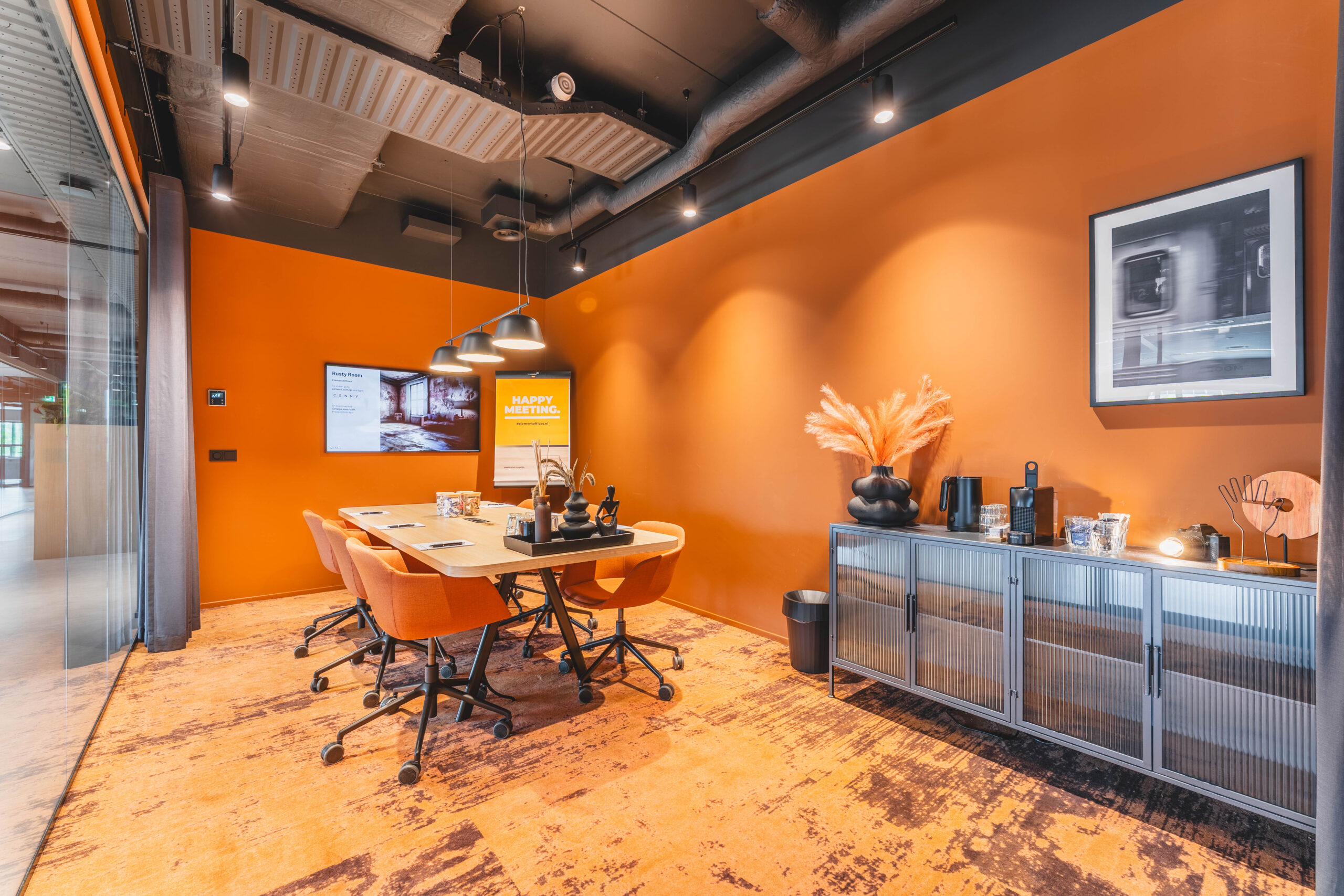 Een moderne vergaderruimte met een oranje thema, voorzien van een tafel, stoelen en een kabinet met decoratie en een koffiezetapparaat.