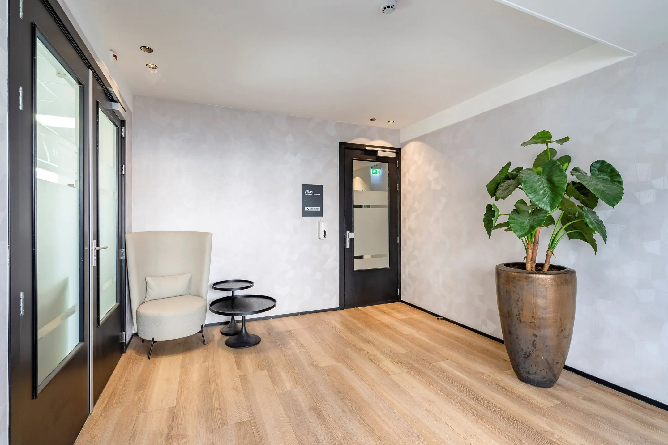 Wachtruimte met een moderne fauteuil, twee bijzettafels, een grote plant in een pot en een deur met het bordje "303_2160".