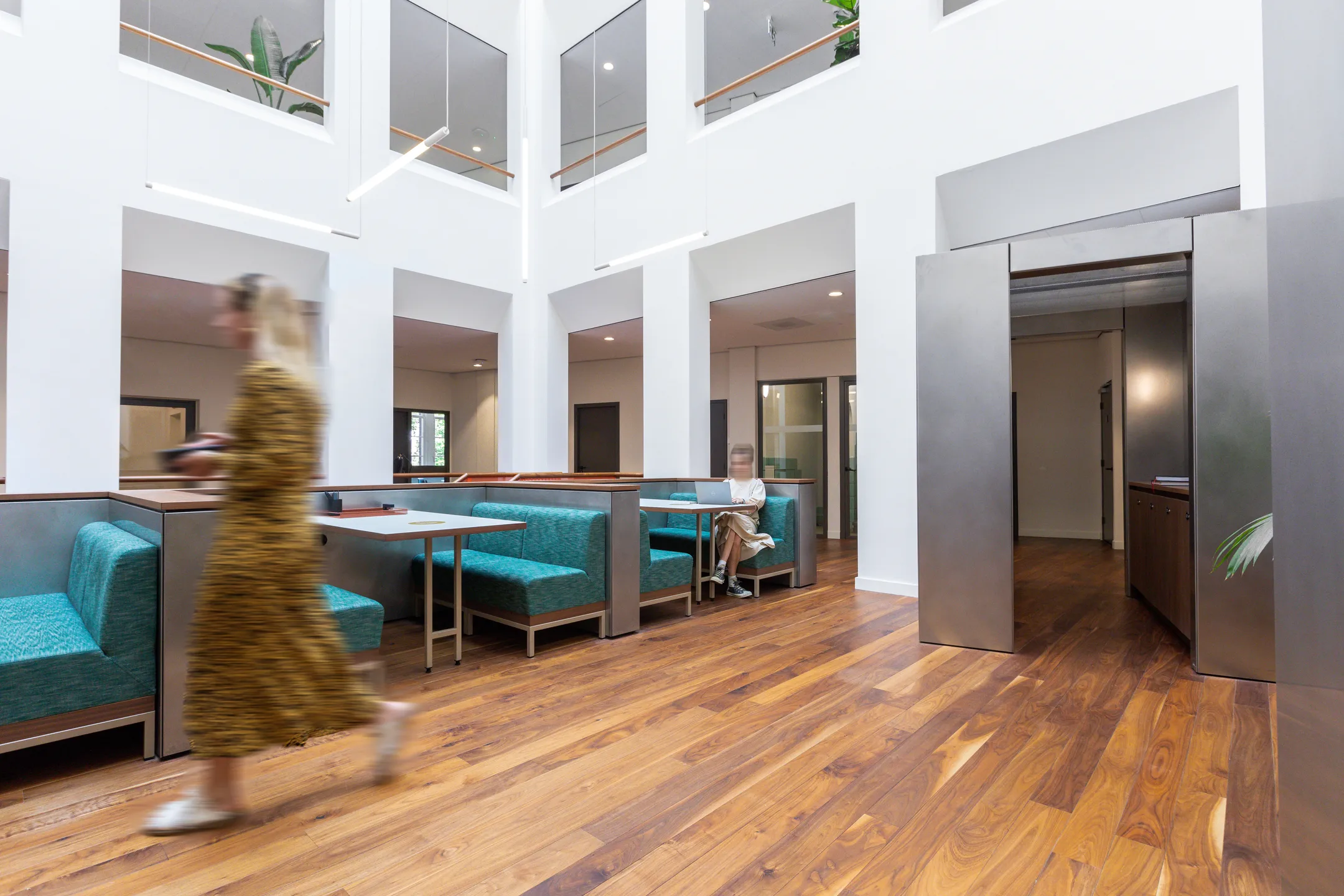 Een moderne en lichte kantoorruimte met een vrouw die voorbij een zitgedeelte loopt en een andere persoon aan een tafel met een laptop zit.