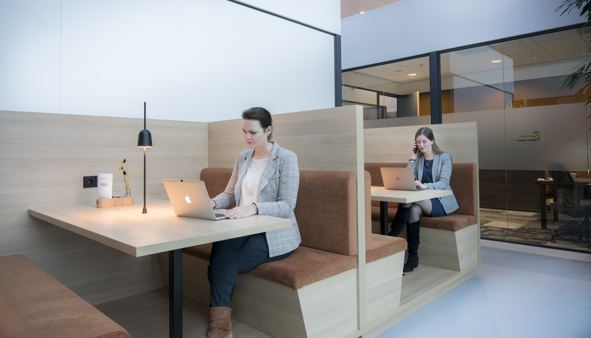 Twee vrouwen werken op laptops in een moderne kantoorruimte met houten treinzitjes.