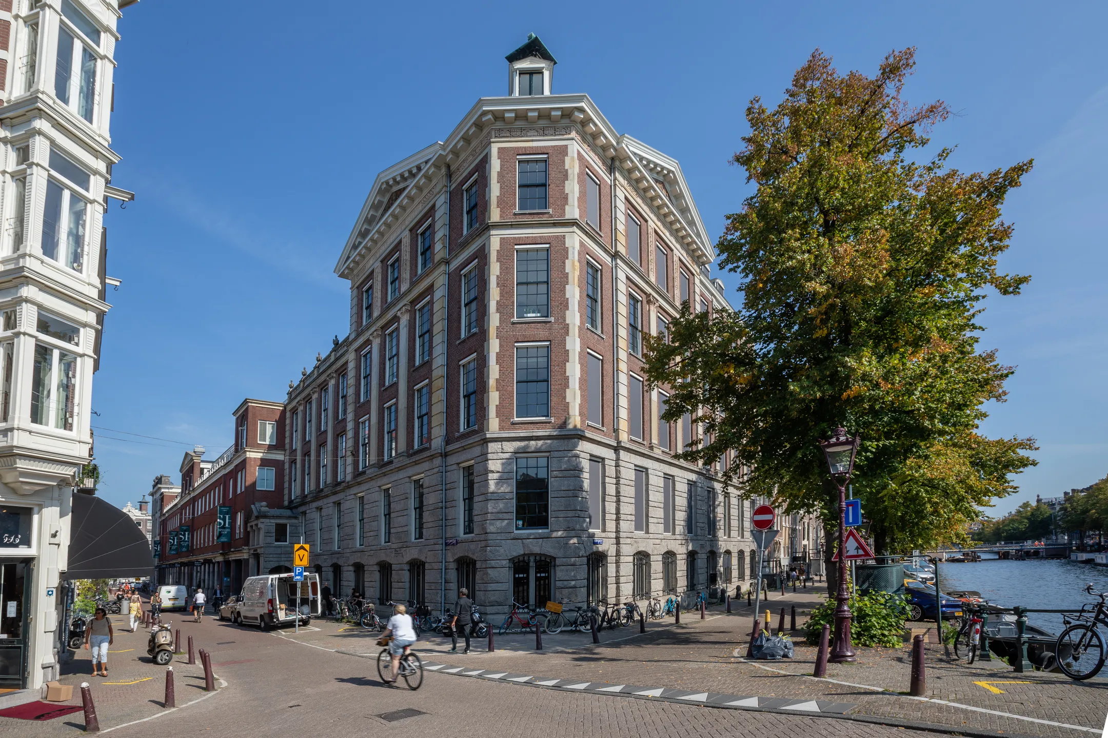 Een zonnige straat in Amsterdam met een monumentaal gebouw aan de gracht en een voorbijfietsende fietser.