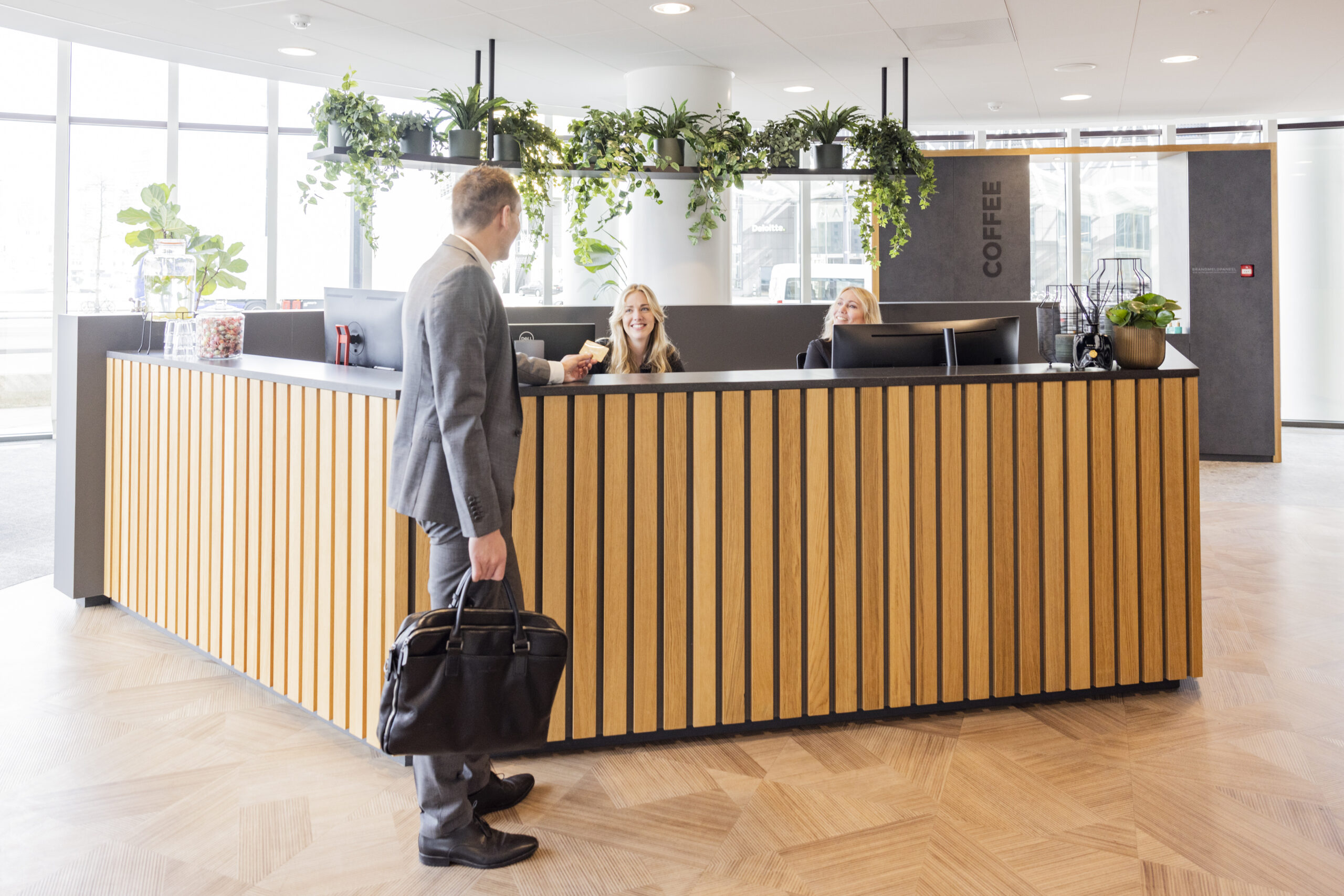Een man in pak ontvangt een kaartje bij een receptie met houten lambrisering en planten eromheen.
