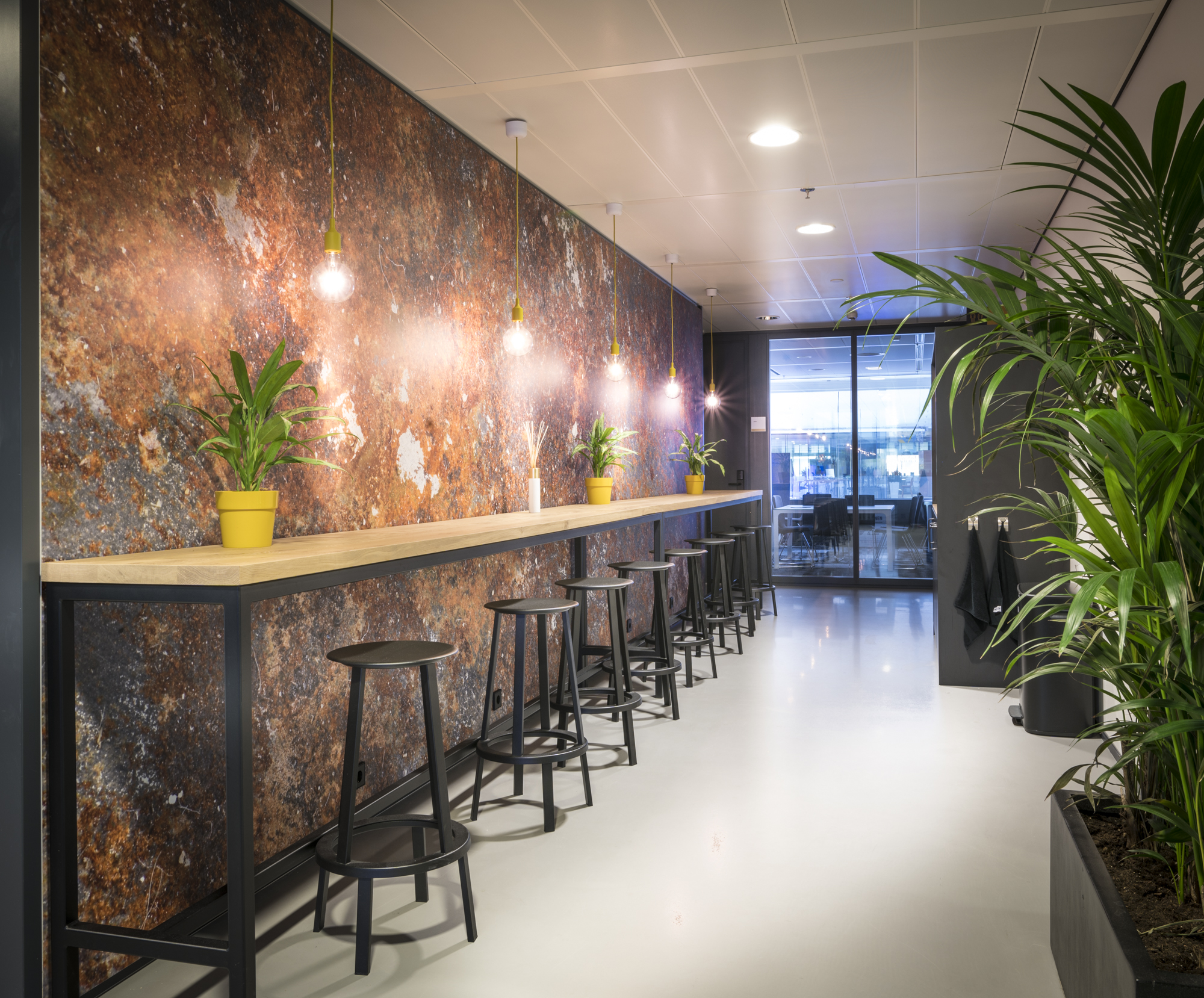Een sfeervolle zithoek met een lange tafel, hoge krukken, planten in gele potten, hanglampen en een glazen deur in een moderne stedelijke ruimte.
