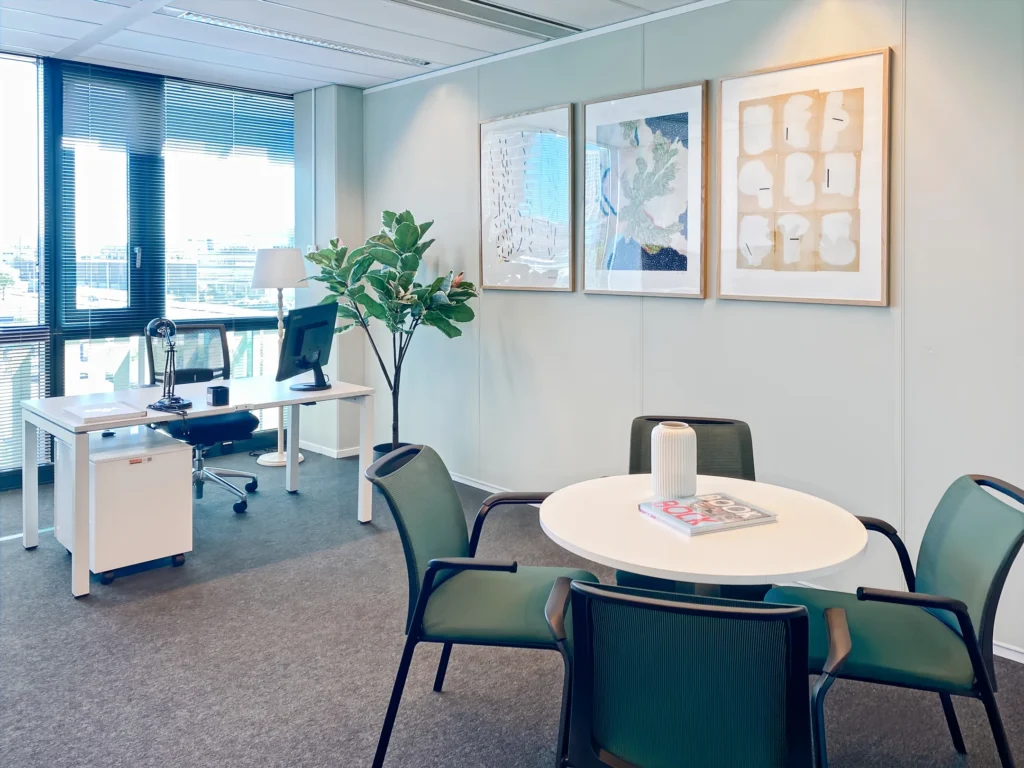 Een modern kantoor met een bureau, een ronde tafel, groene stoelen en kunstwerken aan de muur.