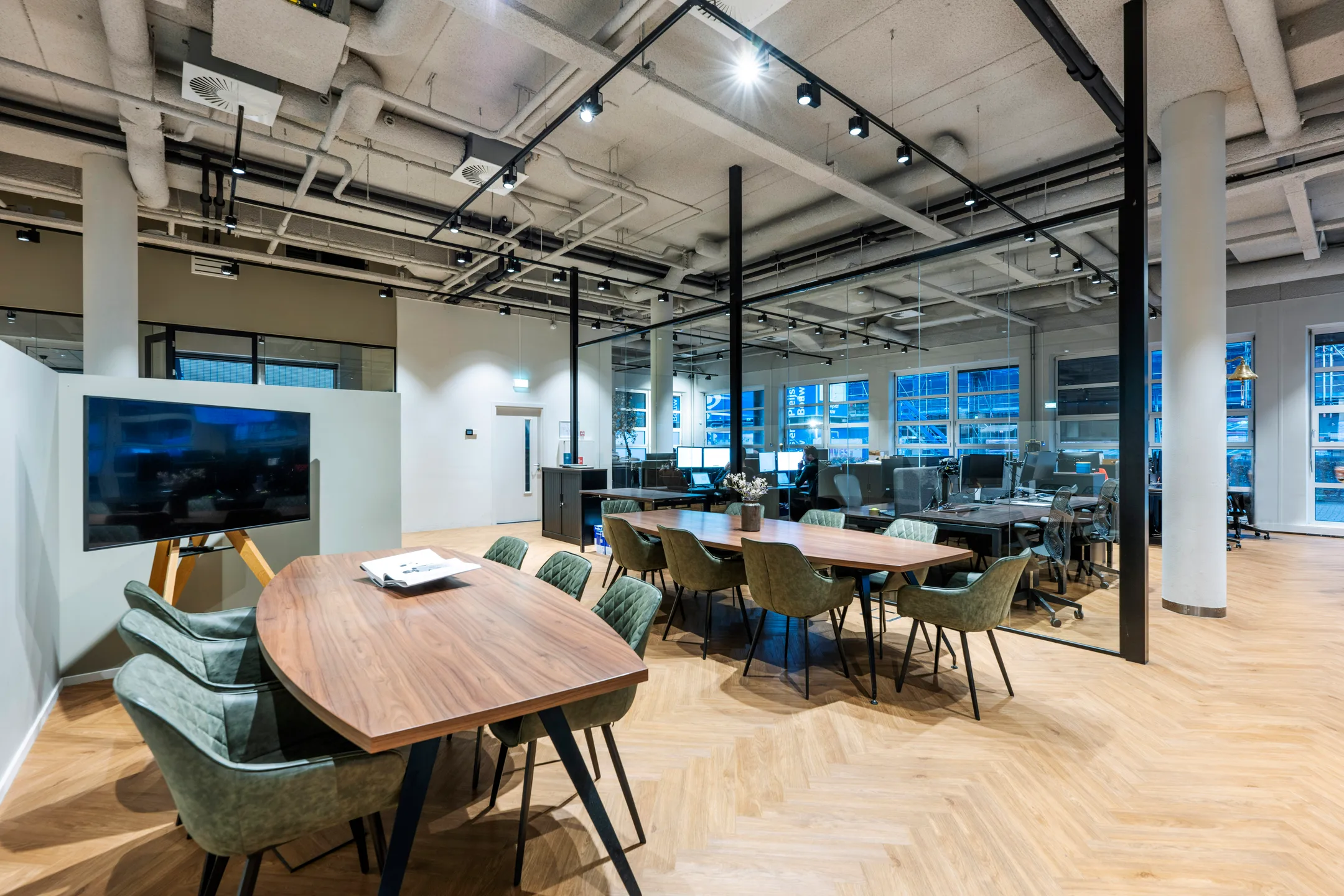 Een moderne kantoorruimte met vergadertafels, groene stoelen en werkplekken onder een plafond met zichtbare leidingen.