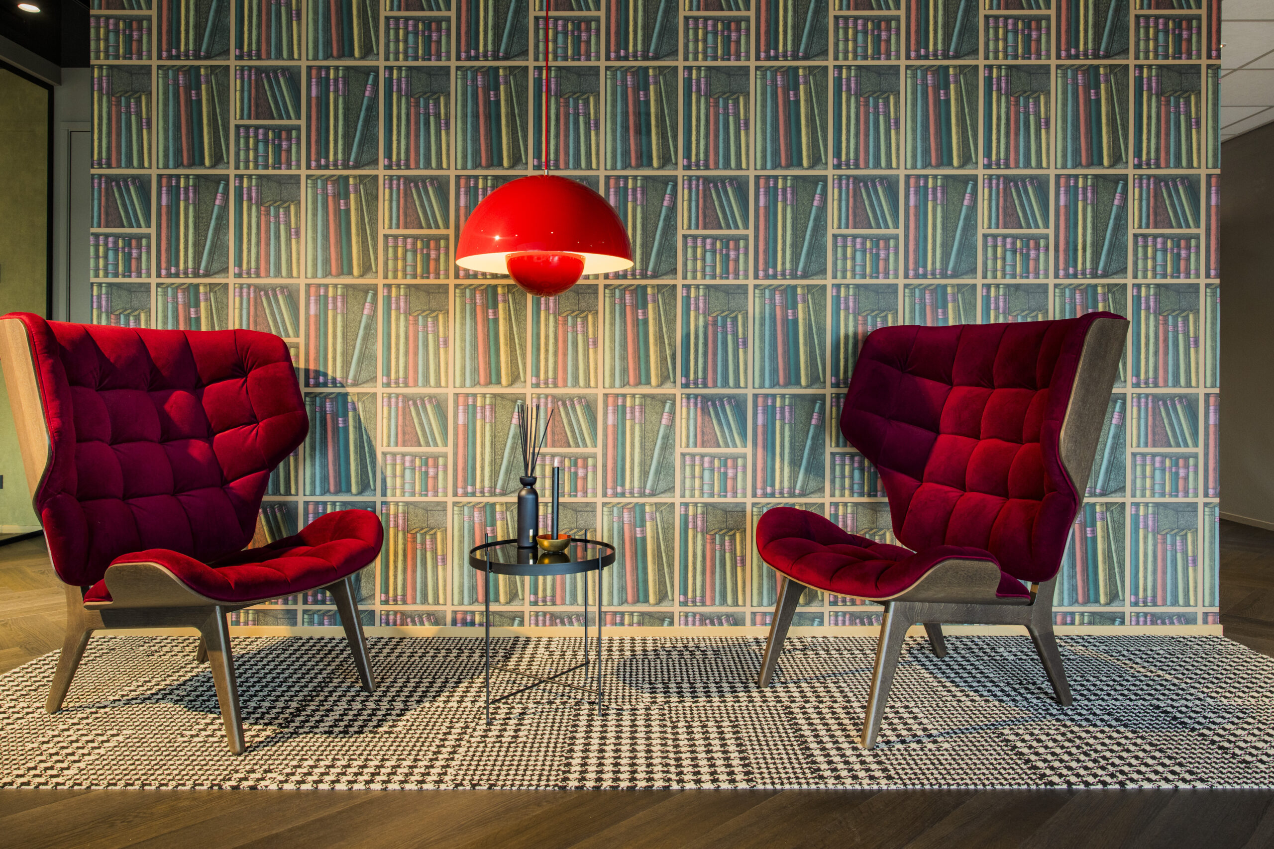Twee rode fauteuils en een zwarte bijzettafel met een moderne lamp tegen een muur met boekenprint behang.
