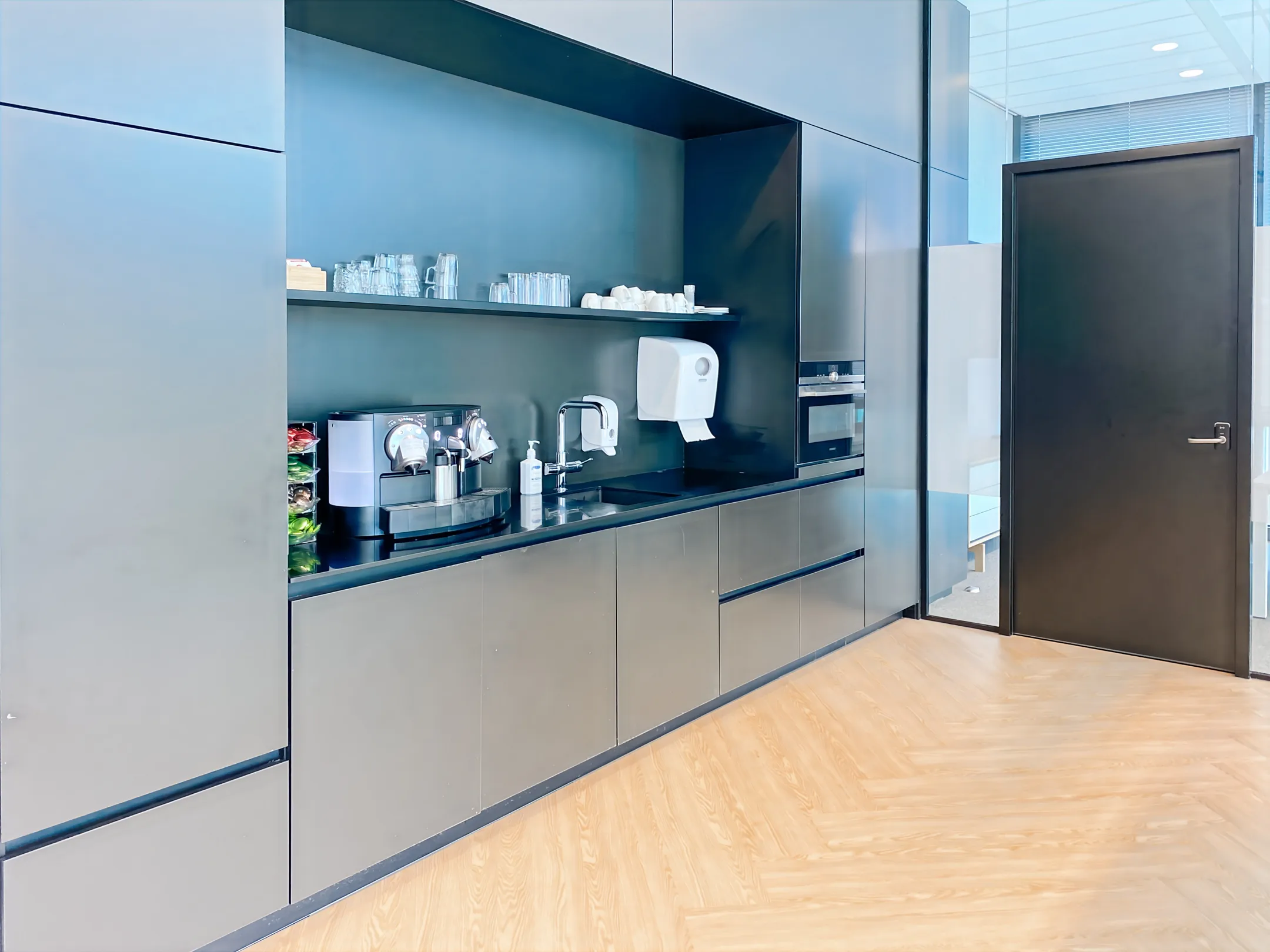 Een moderne kantoor-keuken met een espressoapparaat, een wasbak, glazen en een magnetron in een strakke zwarte kast.