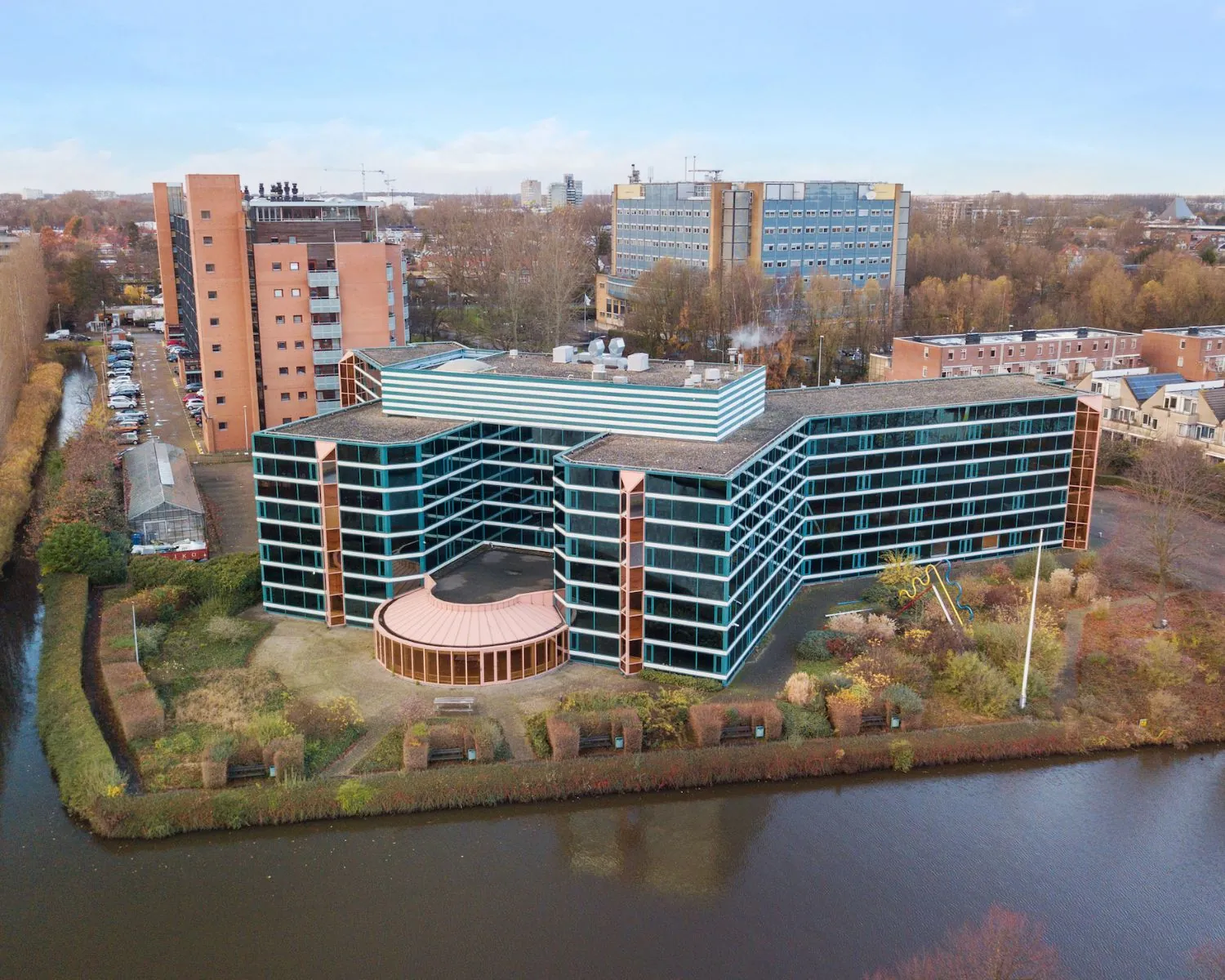 Luchtfoto van gebouw 351 met moderne glazen gevel, gelegen aan een waterpartij en omgeven door woonwijken en parkeerterreinen.