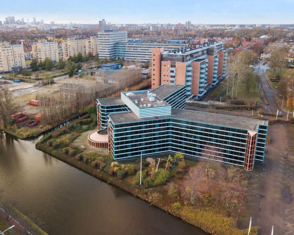 Luchtfoto van een modern kantoorgebouw genaamd 353 met blauwe ramen aan een waterkant, omringd door woongebouwen en bomen.