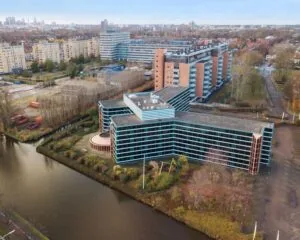 Luchtfoto van een modern kantoorgebouw genaamd 353 met blauwe ramen aan een waterkant, omringd door woongebouwen en bomen.