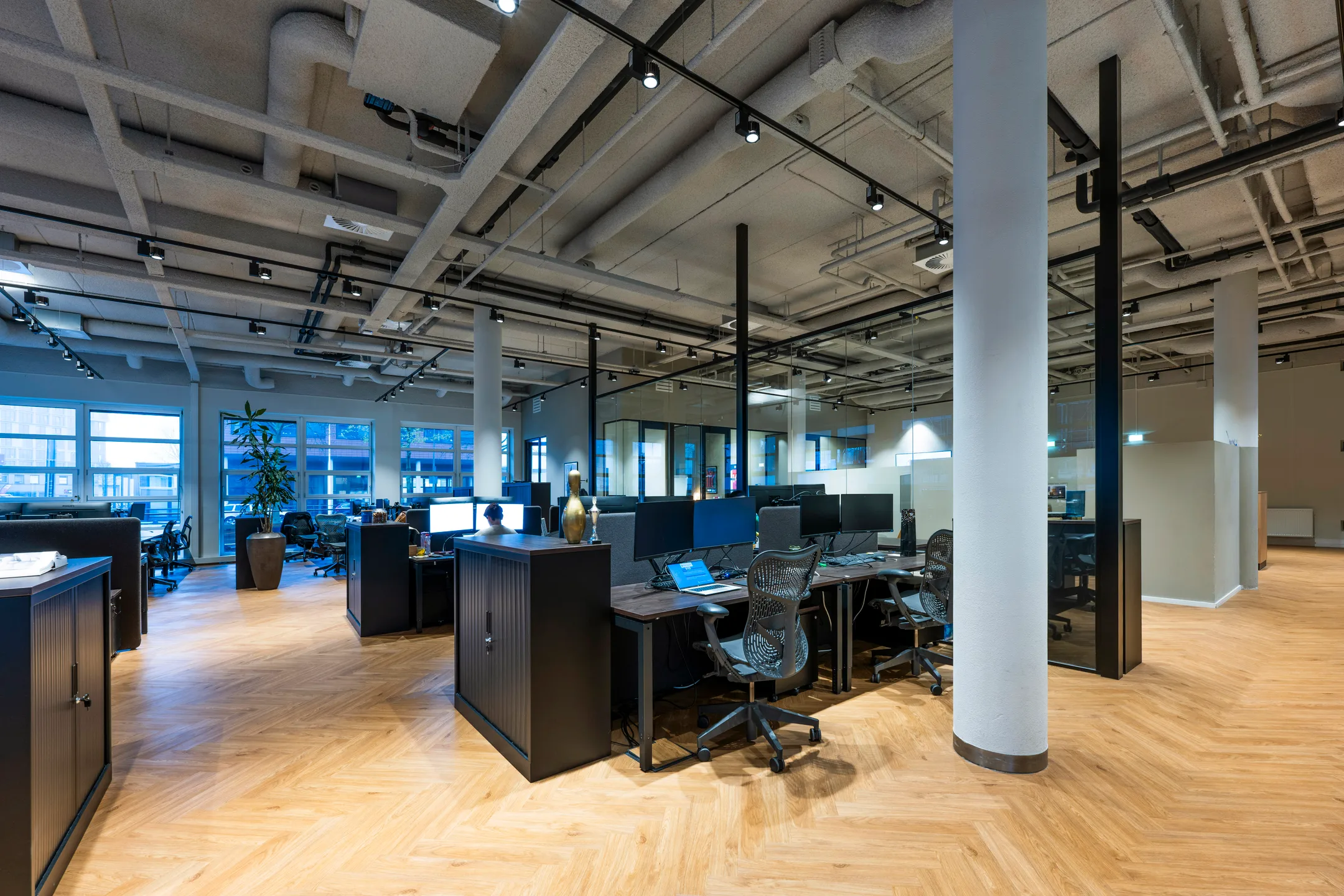 Een moderne, open kantoorruimte met houten vloer en meerdere bureaus en stoelen.