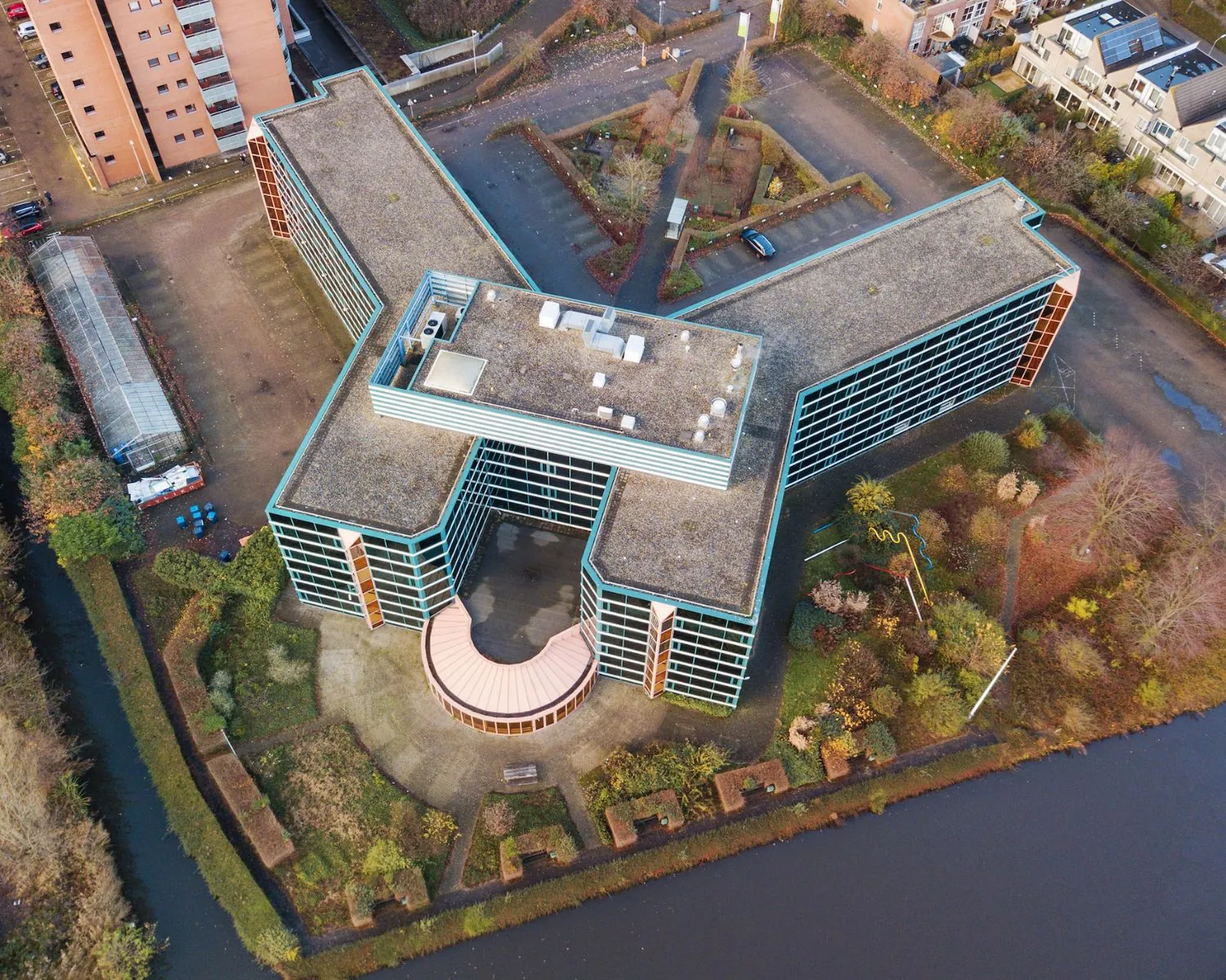 Luchtfoto van een groot kantoorgebouw in H-vorm met een binnenplaats en omliggende parkeerplaatsen, tuinen en water.