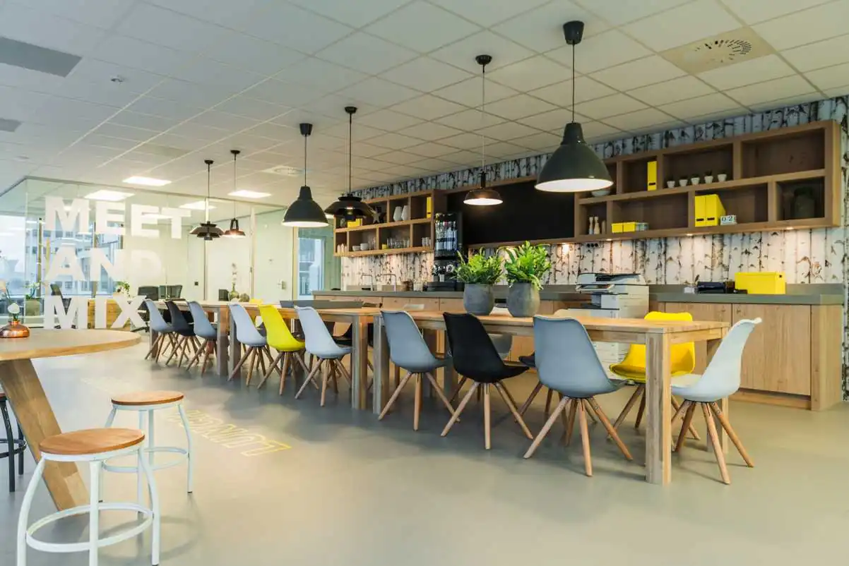Modern ingerichte kantoorlounge op Bisonspoor met lange houten tafels, gekleurde stoelen en een open keukenwand.