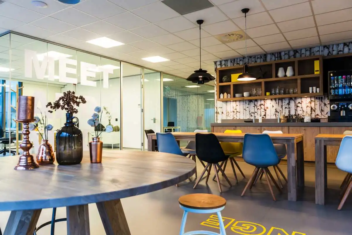 Moderne en kleurrijke kantoorkeuken met houten tafels, diverse stoelen en glazen vergaderruimte met het woord "MEET" op de wand.