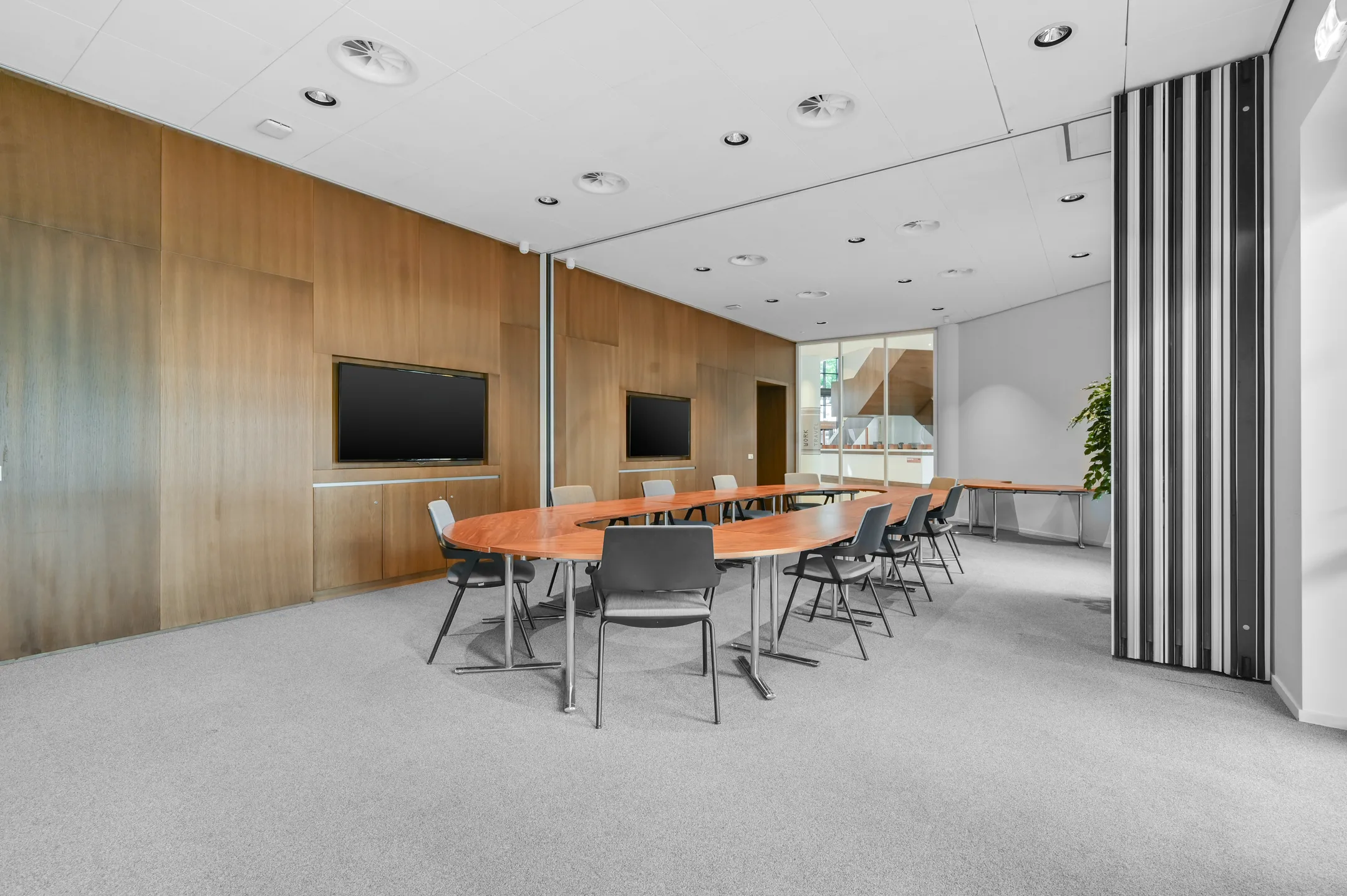 Een moderne vergaderruimte met een ovale houten tafel omringd door zwarte stoelen en twee grote schermen aan de muur.