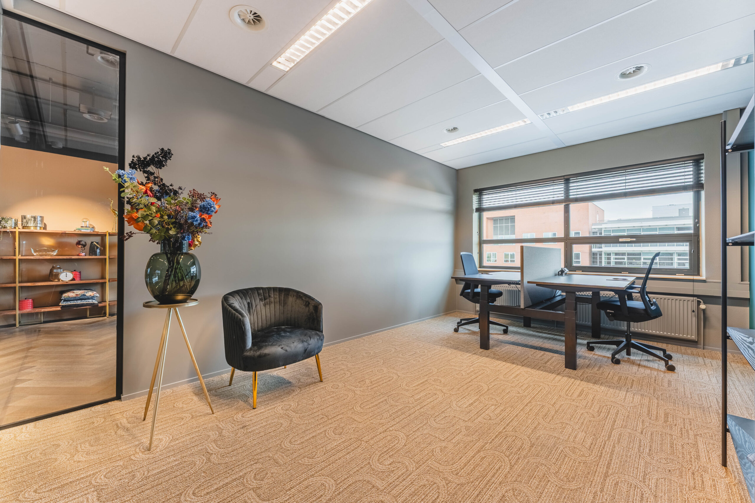 Een modern kantoor met een bureau, twee bureaustoelen, een zwarte stoel en een vaas met kleurrijke bloemen.