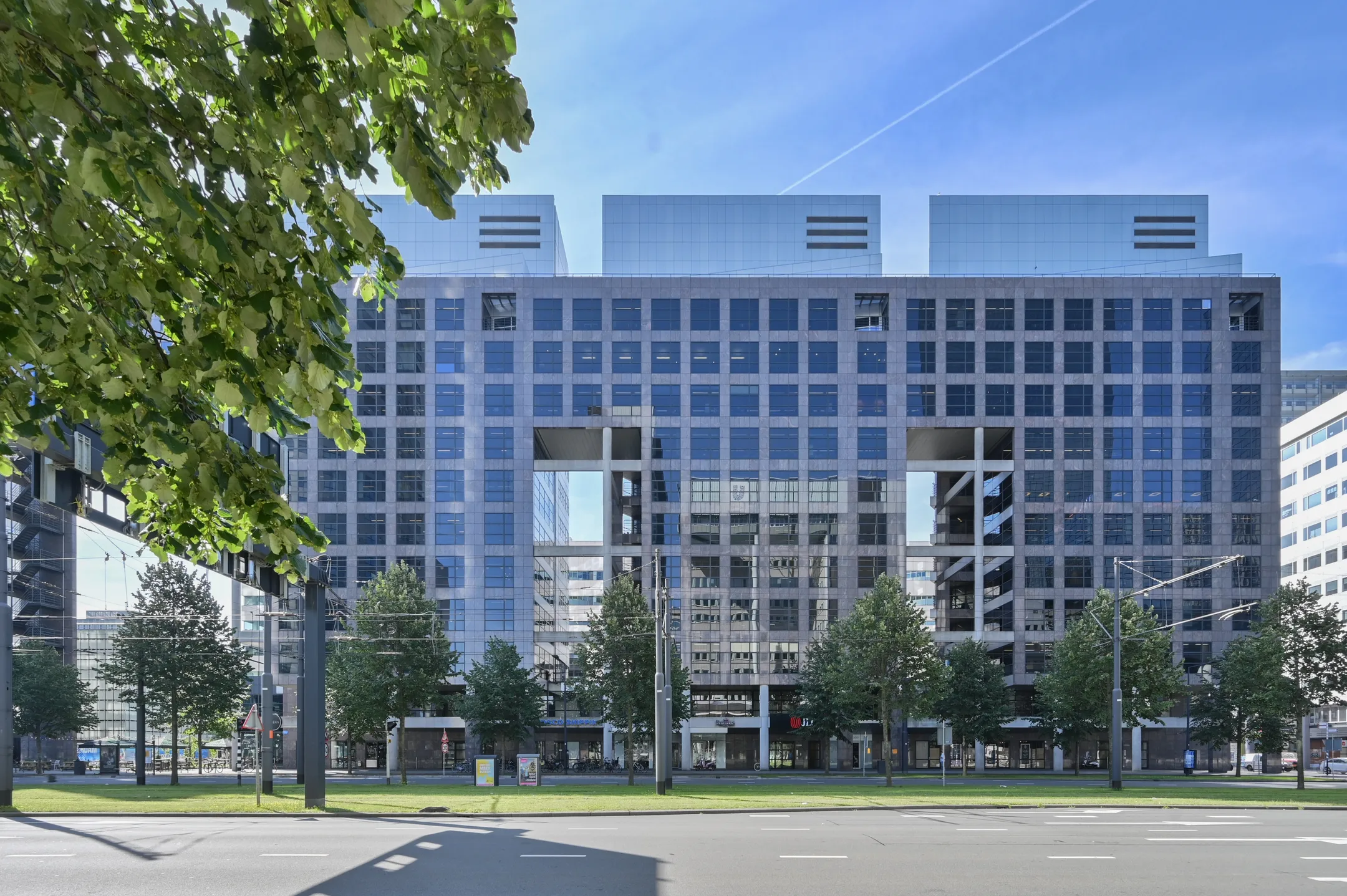 Een modern kantoorgebouw met een glazen gevel en groene bomen op de voorgrond.