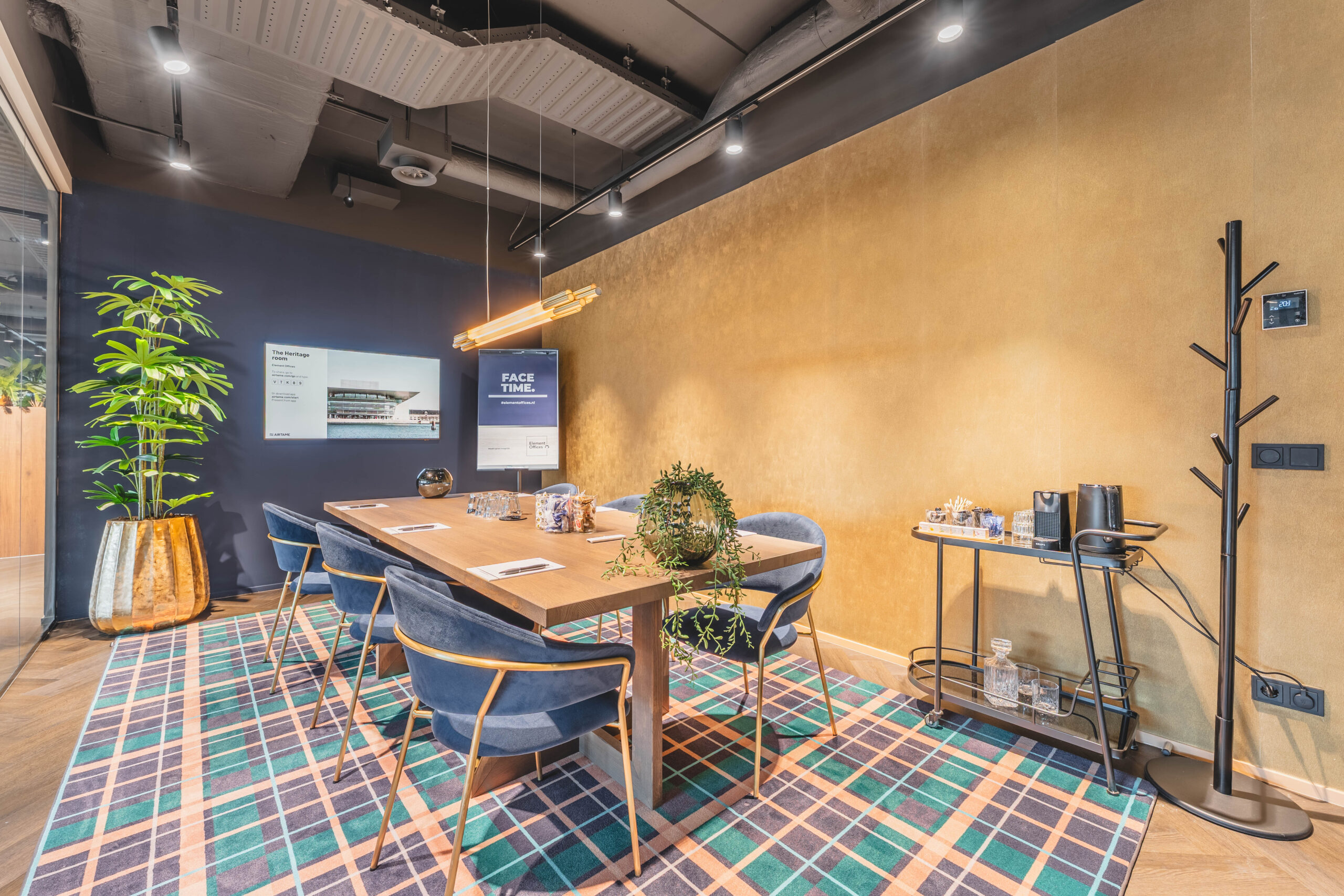 Een moderne vergaderzaal met een lange tafel, blauwe stoelen, groene plant en warme verlichting.