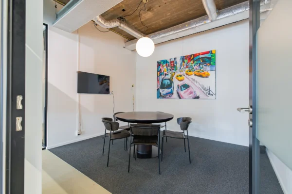 Een vergaderruimte met een ronde tafel en stoelen, een kleurrijk schilderij aan de muur en een televisie.