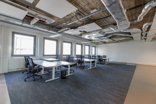 Een moderne, open kantoorruimte met bureaus en bureaustoelen, met zichtbare leidingen aan het plafond.