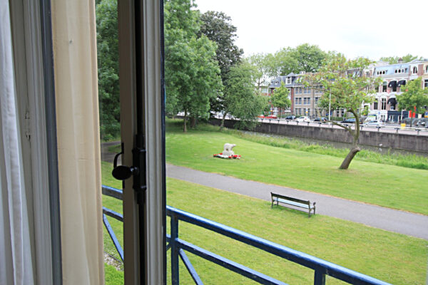 Uitzicht vanuit een open raam op de Museumlaan met een grasveld, beeldhouwwerk, bankje en woonhuizen op de achtergrond.