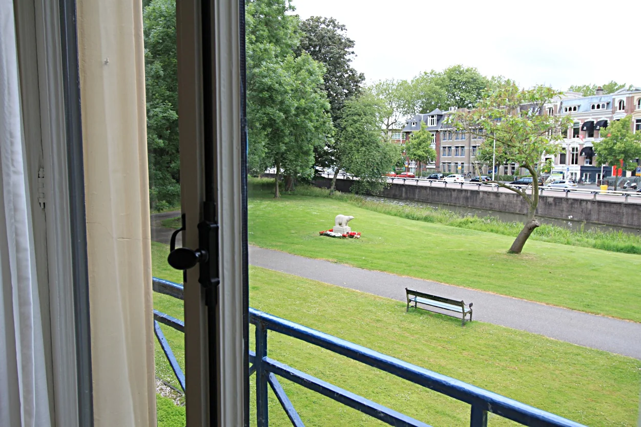 Uitzicht vanuit een open raam op de Museumlaan met een grasveld, beeldhouwwerk, bankje en woonhuizen op de achtergrond.