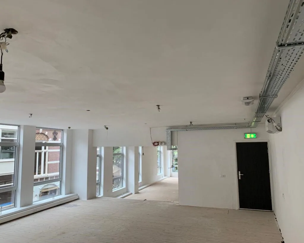 Een lege, lichtgevende ruimte in verbouwing met grote ramen en zicht op een nooduitgangsdeur.
