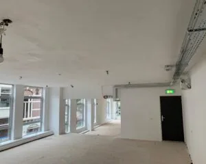 Een lege, lichtgevende ruimte in verbouwing met grote ramen en zicht op een nooduitgangsdeur.