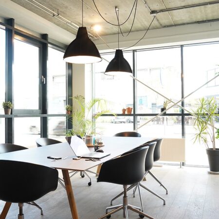 Een moderne vergaderruimte met een grote tafel, zwarte stoelen, hanglampen en veel natuurlijk licht.