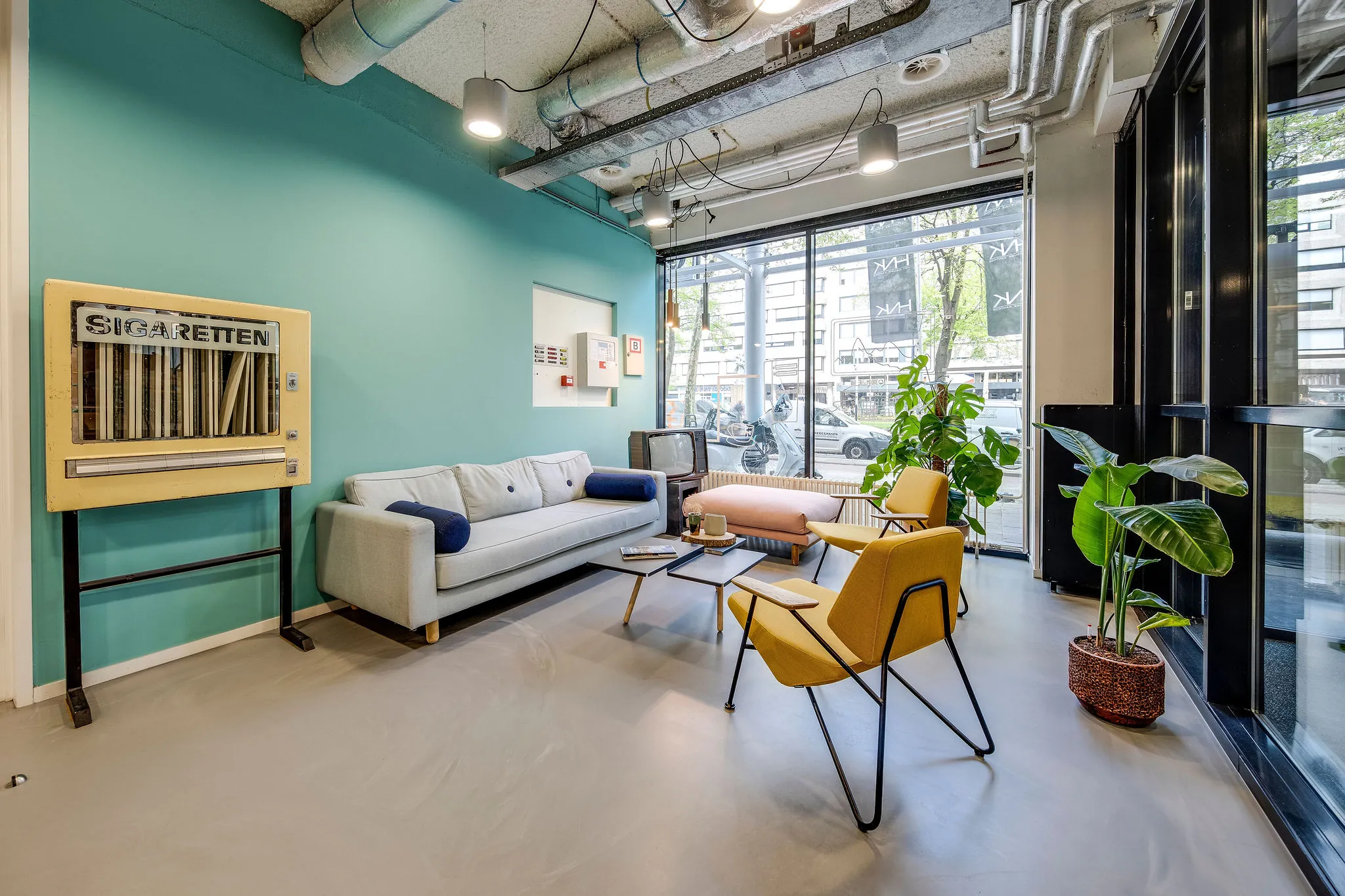 Moderne loungehoek in HNK Rotterdam Centrum met vintage sigarettenautomaat, zitbanken en kamerplanten.