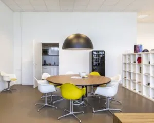 Moderne kantoorruimte aan de Panamalaan 104 in Amsterdam met een ovale vergadertafel, gele en witte stoelen, een grote hanglamp en een open keuken op de achtergrond.