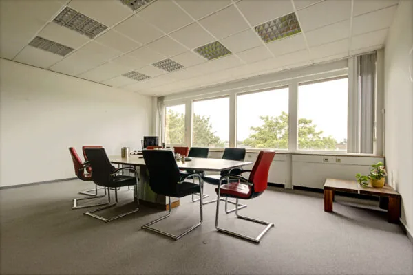 Vergaderruimte aan de Maarssenbroeksedijk met grote tafel, zes stoelen en uitzicht op bomen door brede ramen.