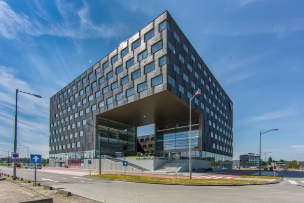 Een modern, kubusvormig gebouw met glazen entree en omliggende straten onder een blauwe hemel.