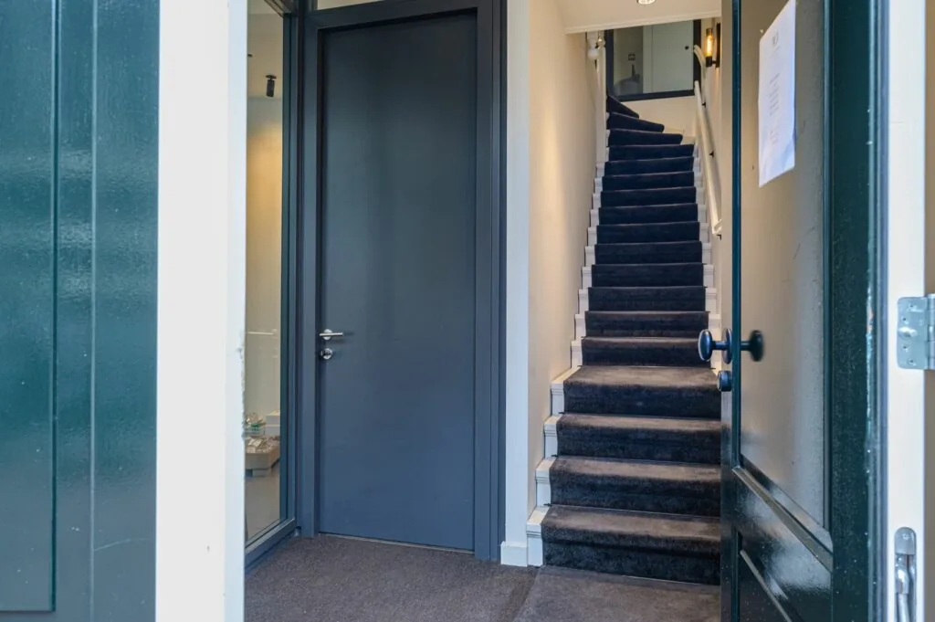 Openstaande deur met zicht op een trap bekleed met donker tapijt en een gesloten grijze binnendeur links.
