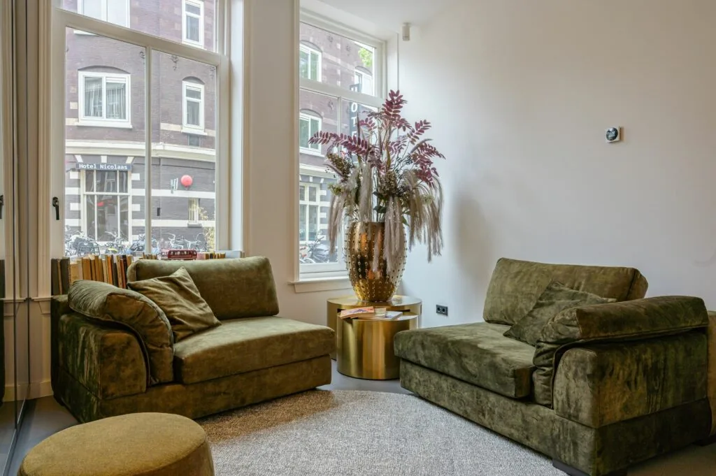 Stijlvol ingerichte zithoek met twee groene fluwelen fauteuils, een gouden bijzettafel en een sierlijke vaas met droogbloemen, met uitzicht op Hotel Nicolaas.
