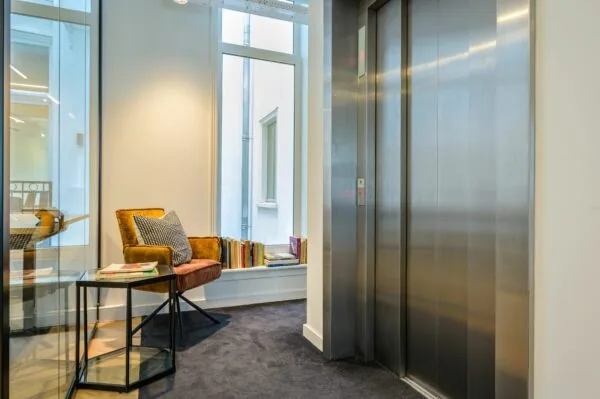 Moderne wachtruimte met een comfortabele stoel, boeken en een glazen bijzettafel naast een gesloten liftdeur.