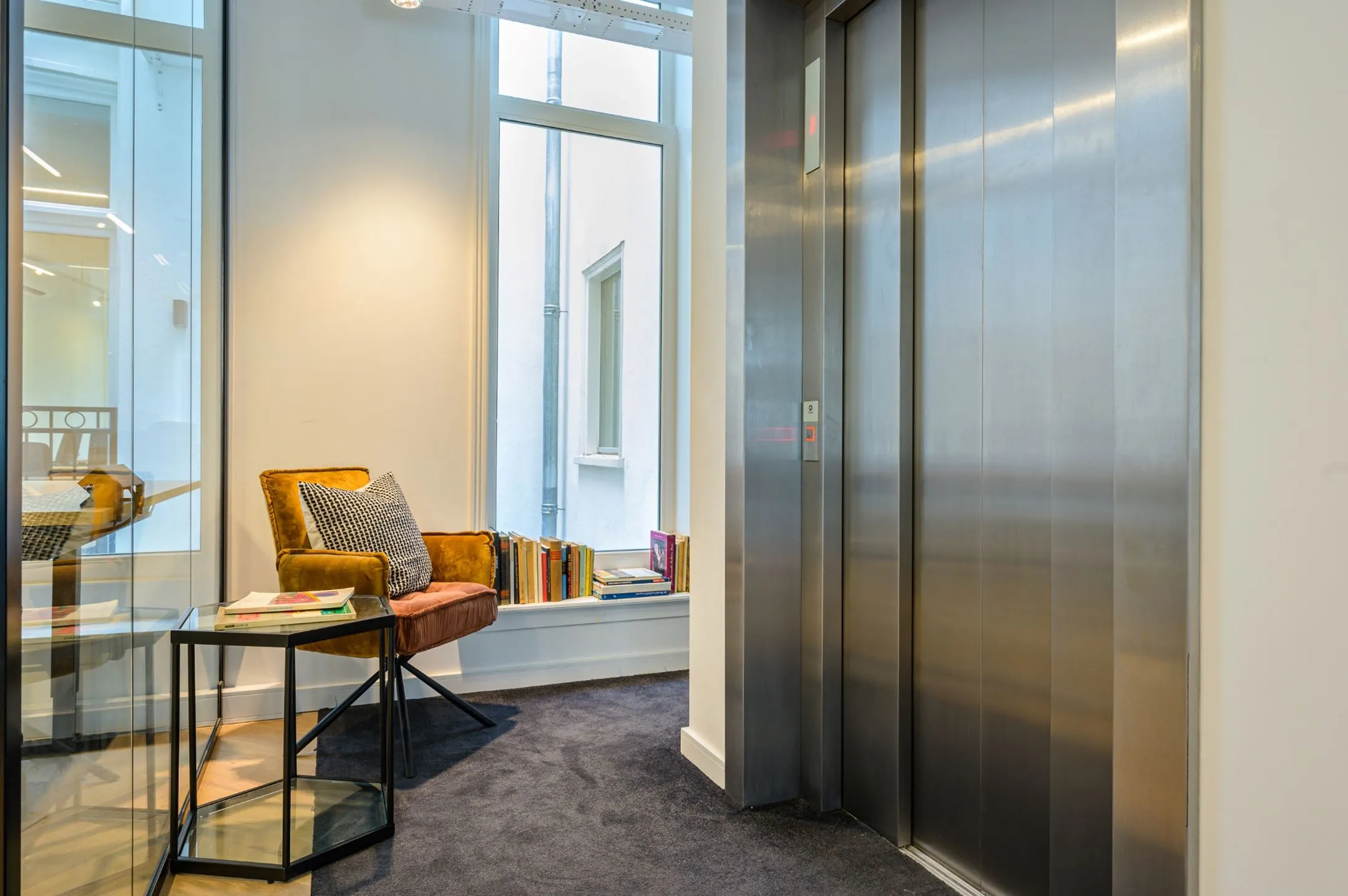 Moderne wachtruimte met een comfortabele stoel, boeken en een glazen bijzettafel naast een gesloten liftdeur.
