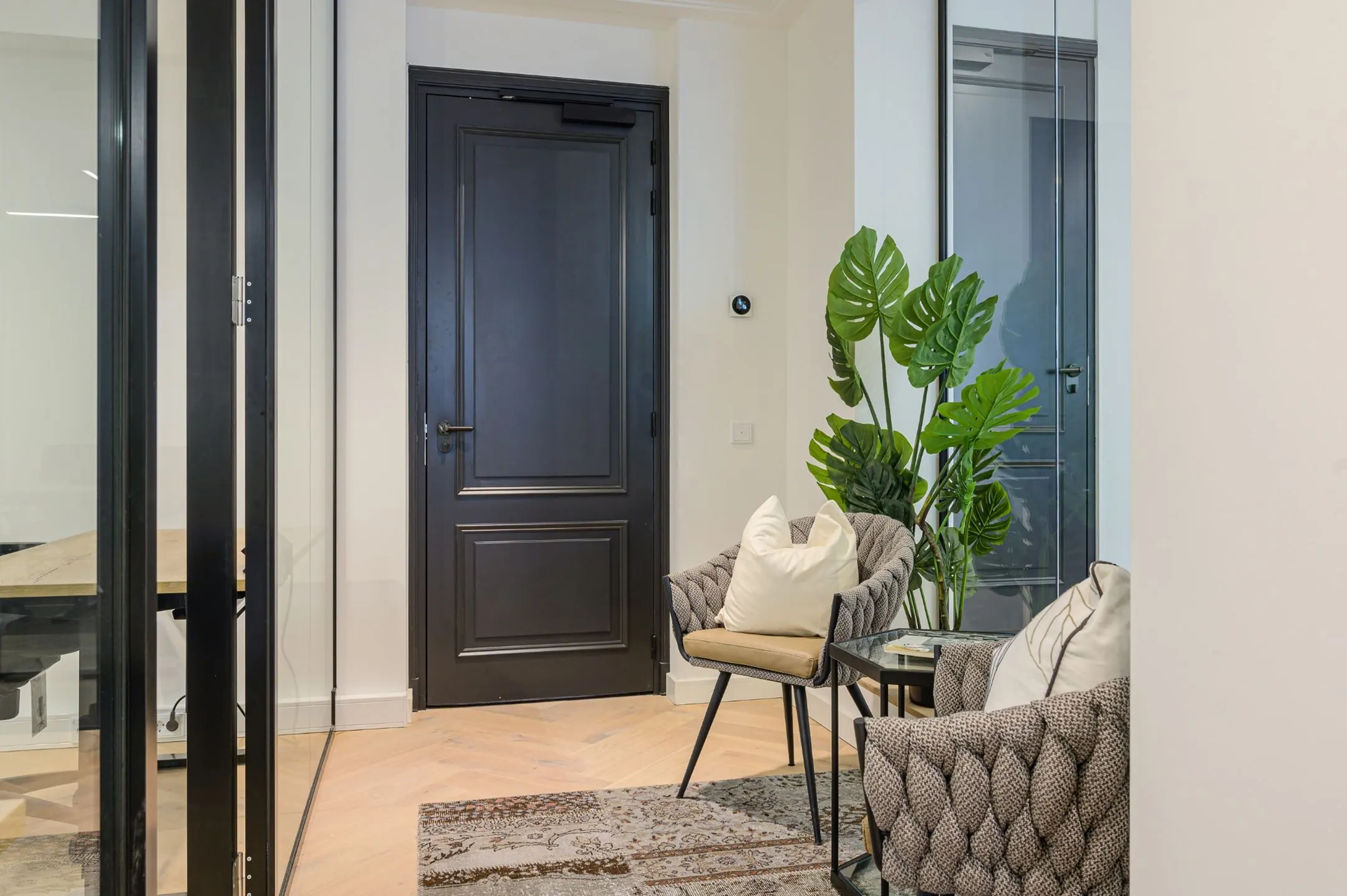 Moderne wachtruimte met twee gestoffeerde stoelen, een glazen bijzettafel en grote groene kamerplant naast een zwarte deur.