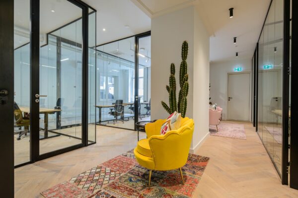 Moderne kantoorruimte met glazen wanden, een gele fauteuil op een kleurrijk vloerkleed en een hoge cactus tegen de muur.