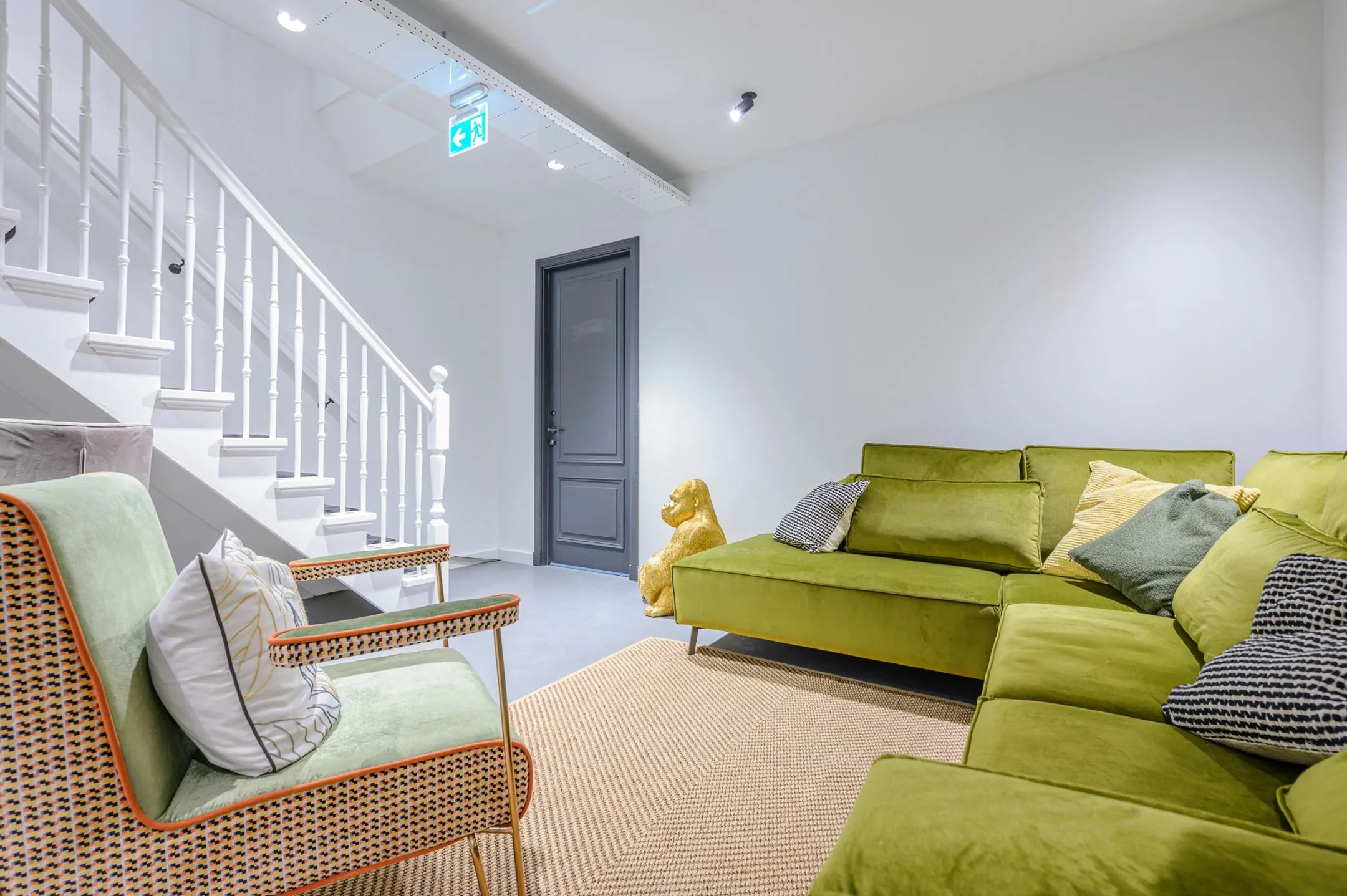 Moderne loungehoek met groene fluwelen bank, designstoel, beige vloerkleed en witte trap in een strak interieur.