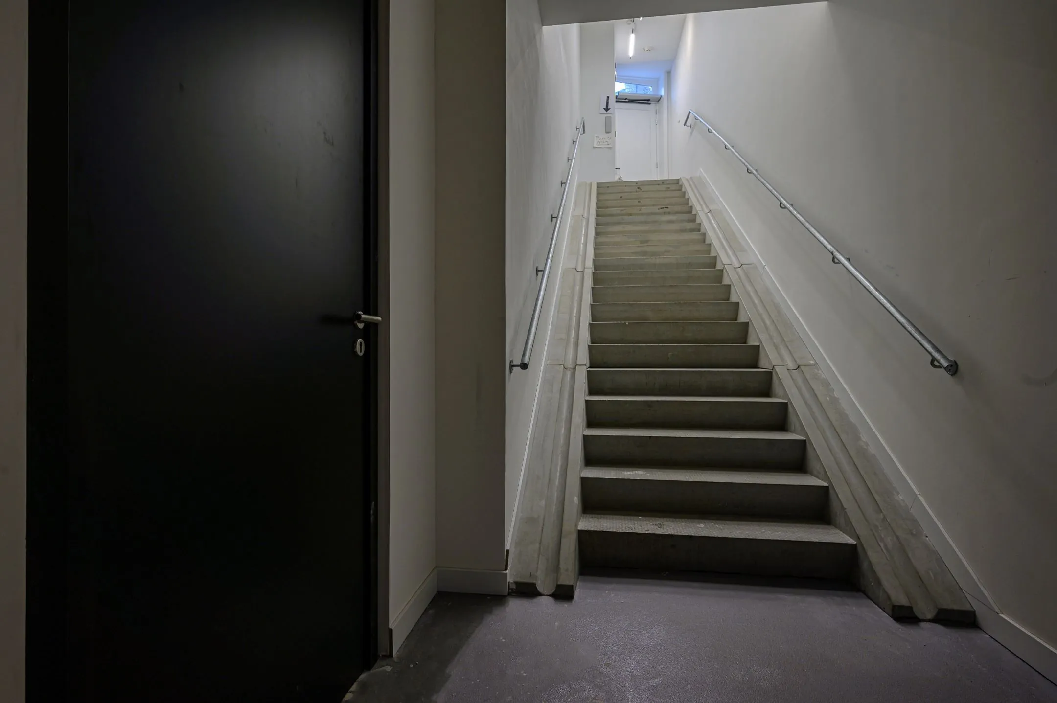 Betonnen trap met metalen leuningen in een smalle, grijze gang met een zwarte deur aan de linkerkant.