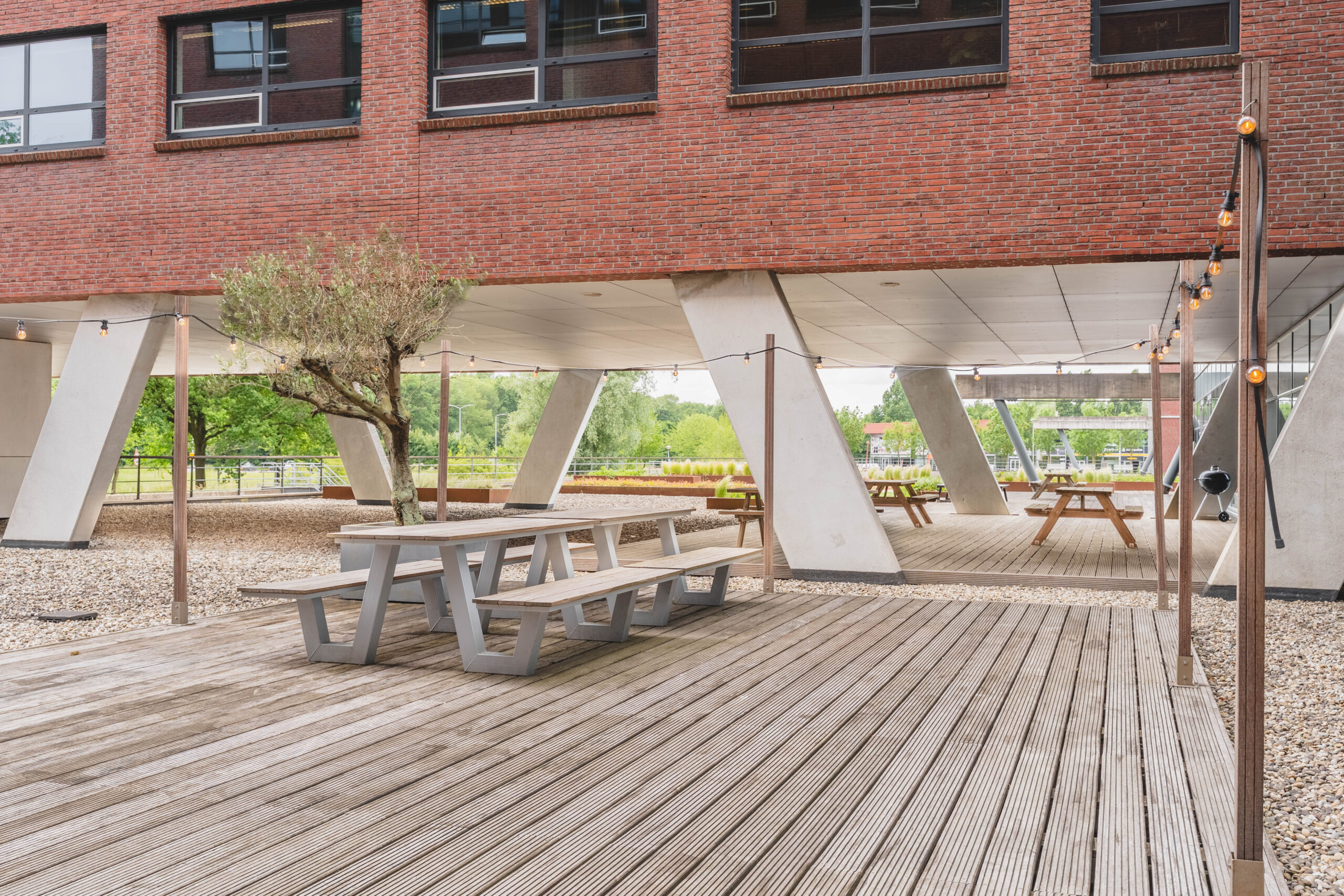 Terras bij Rijnzathe met houten picknicktafels en sfeerverlichting onder een modern gebouw.