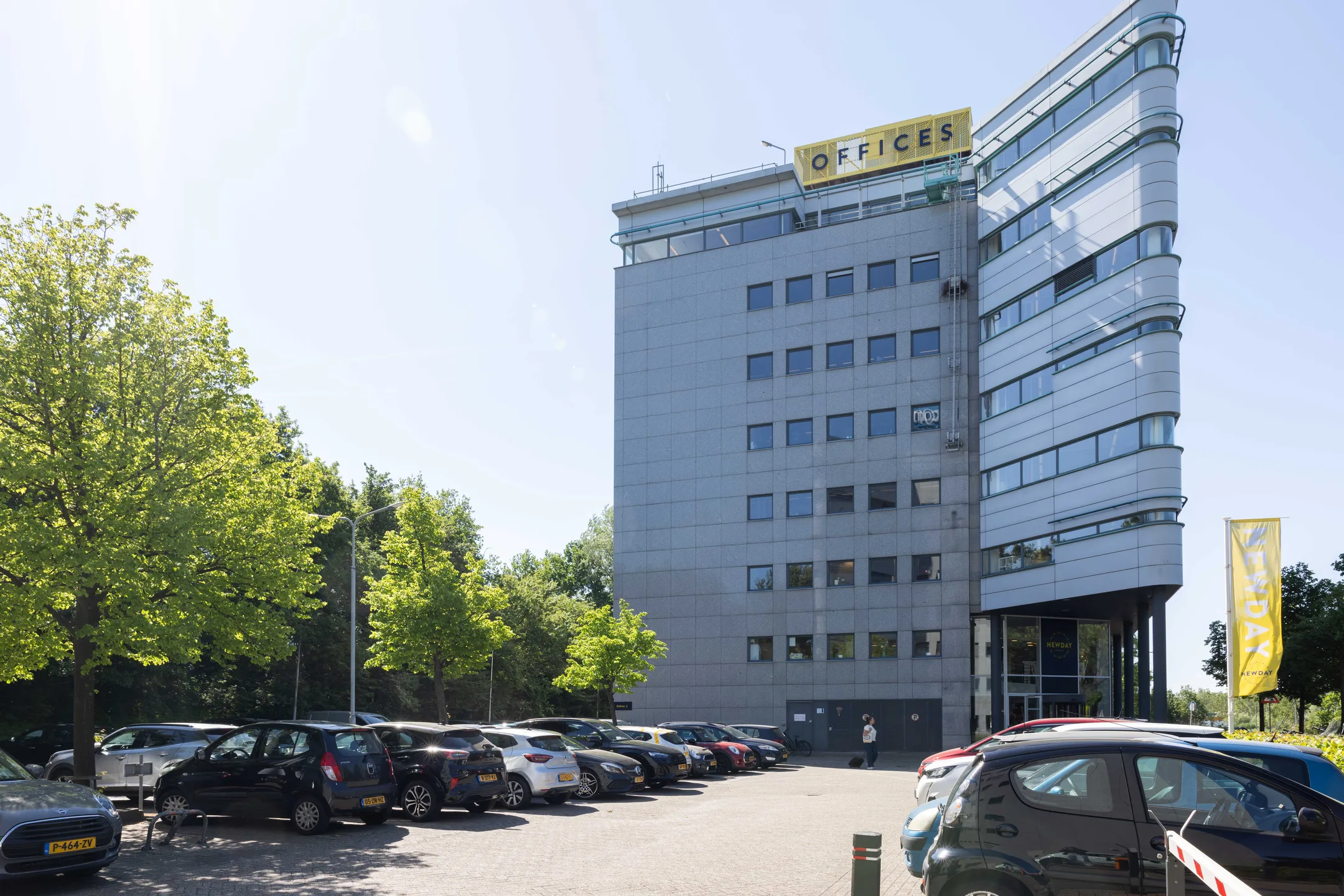 Een modern kantoorgebouw met parkeerplaats en groene bomen ervoor.