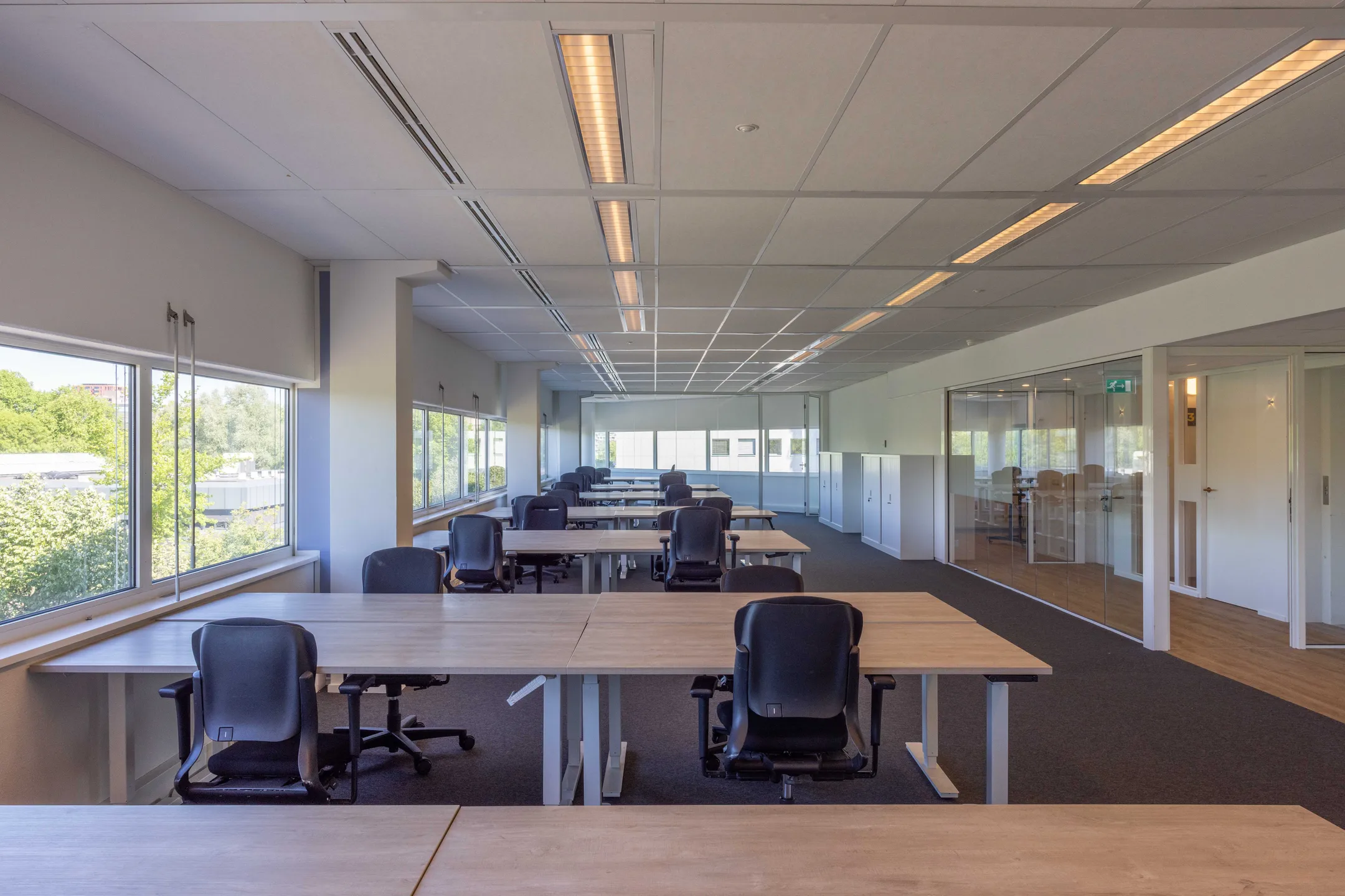 Een moderne, lege kantoorruimte met bureaus en stoelen, verlicht door natuurlijk licht van grote ramen.