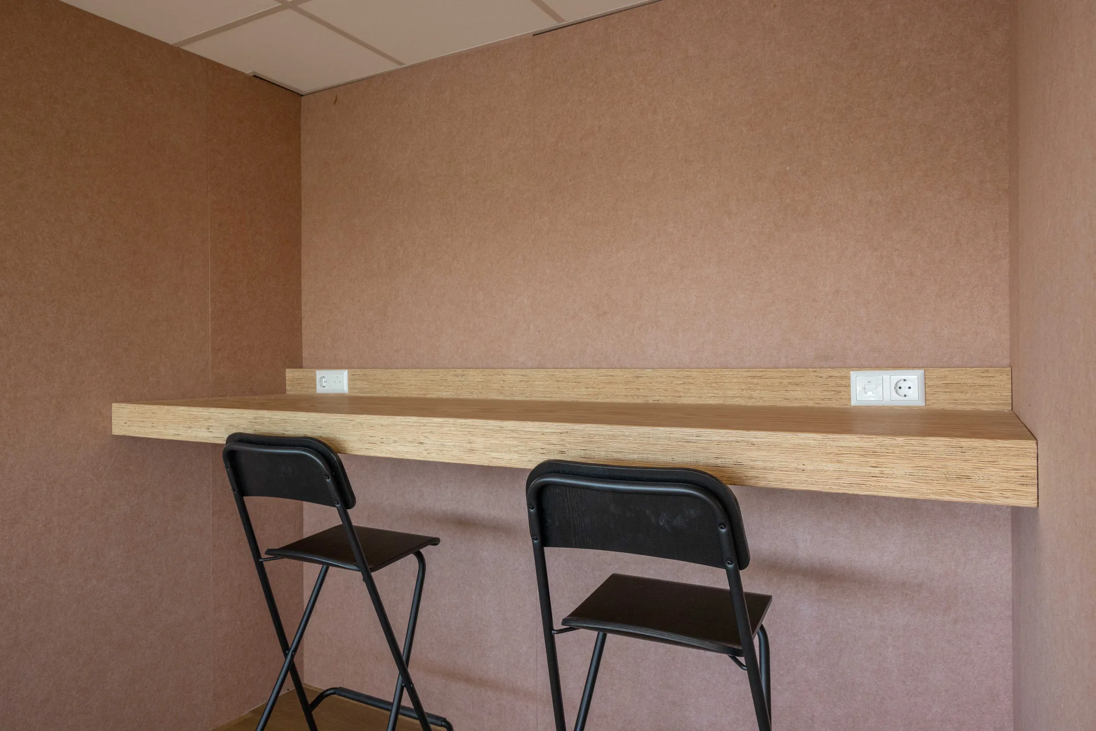 Twee zwarte klapstoelen staan voor een houten werkblad met stopcontacten aan een beige muur.