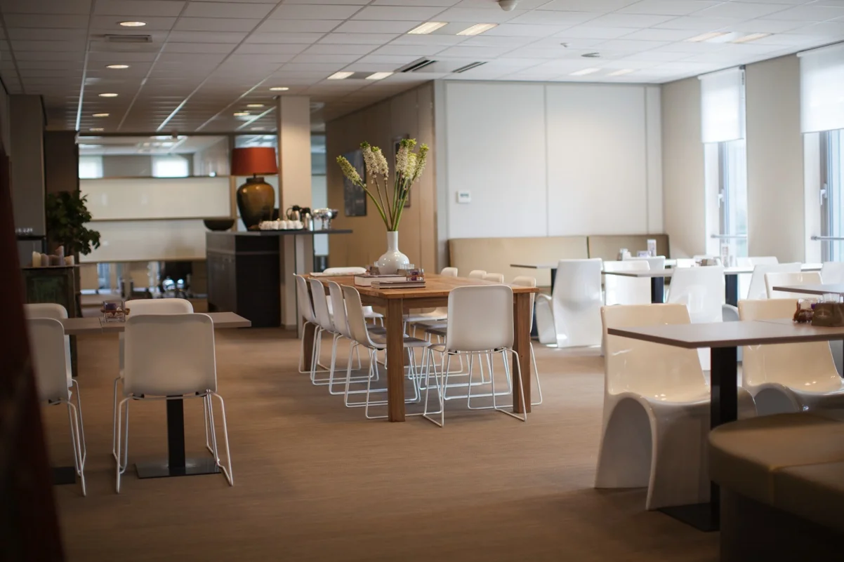Moderne en netjes ingerichte bedrijfs- of kantoorkantine aan de Orteliuslaan met houten tafels, witte stoelen en een grote vaas met bloemen.