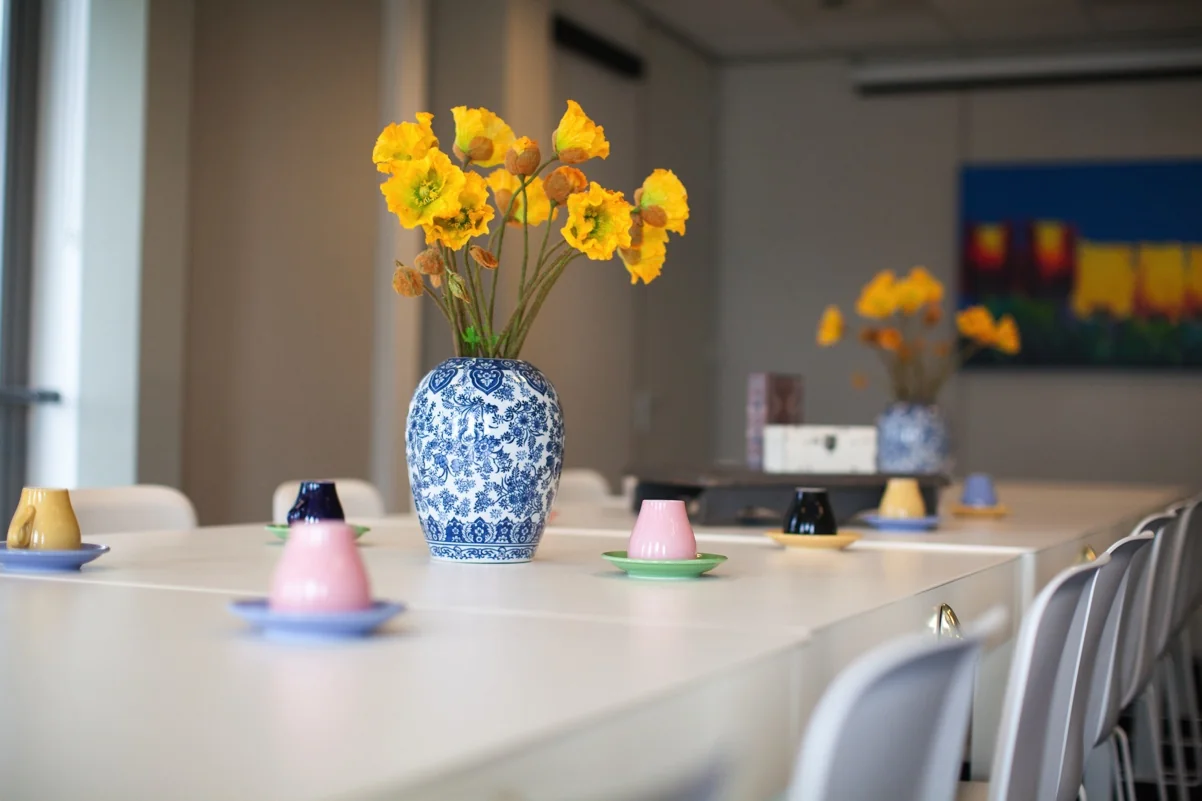 Vergaderruimte aan de Orteliuslaan met een lange tafel, felgekleurde kopjes en een grote blauwe vaas met gele bloemen.