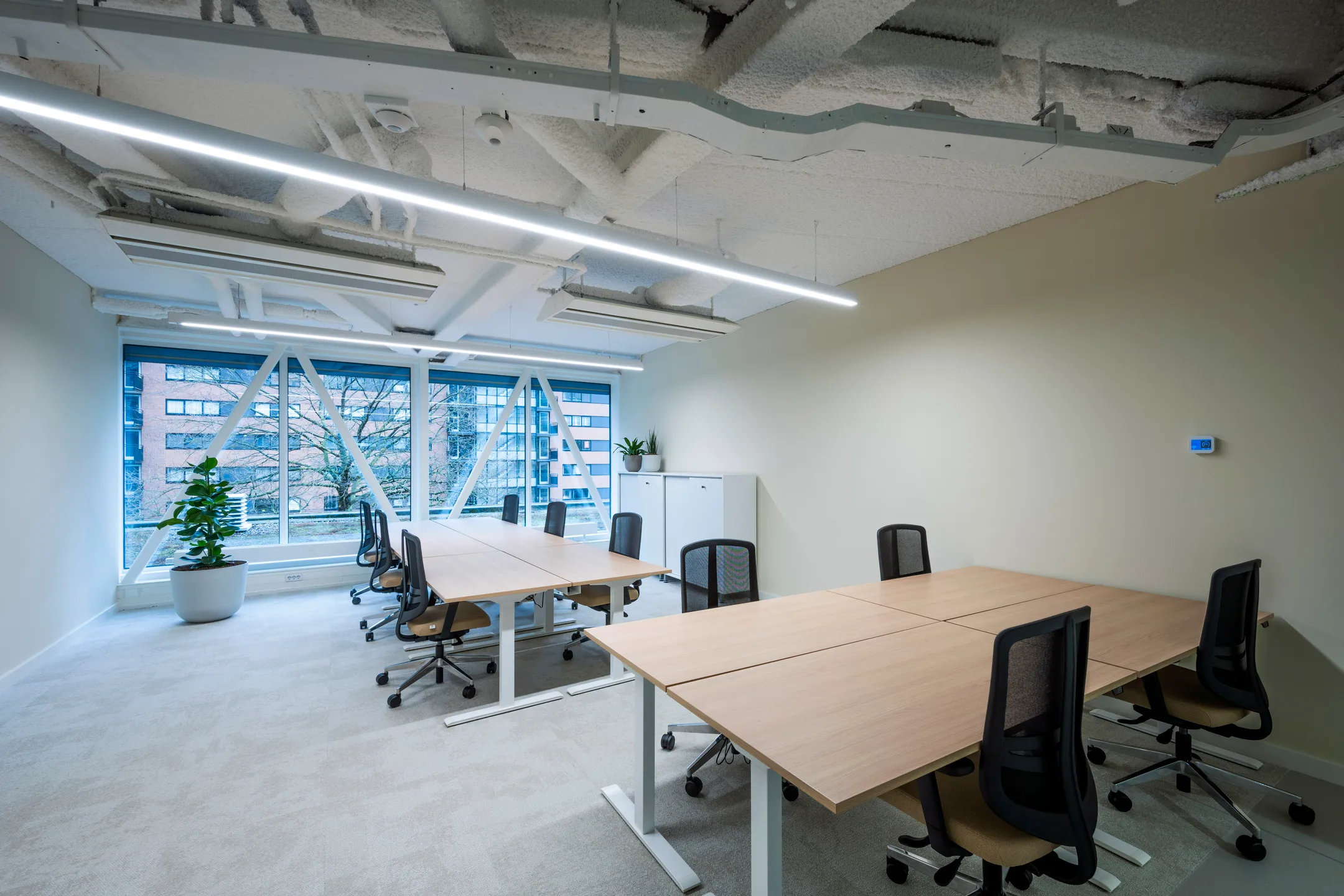 Een moderne kantoorruimte met bureaus, bureaustoelen en grote ramen met uitzicht op een gebouw.