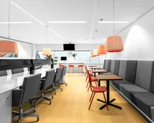 Moderne kantoorruimte met lange werktafels, grijze stoelen en een zitgedeelte met rode accenten en hanglampen.