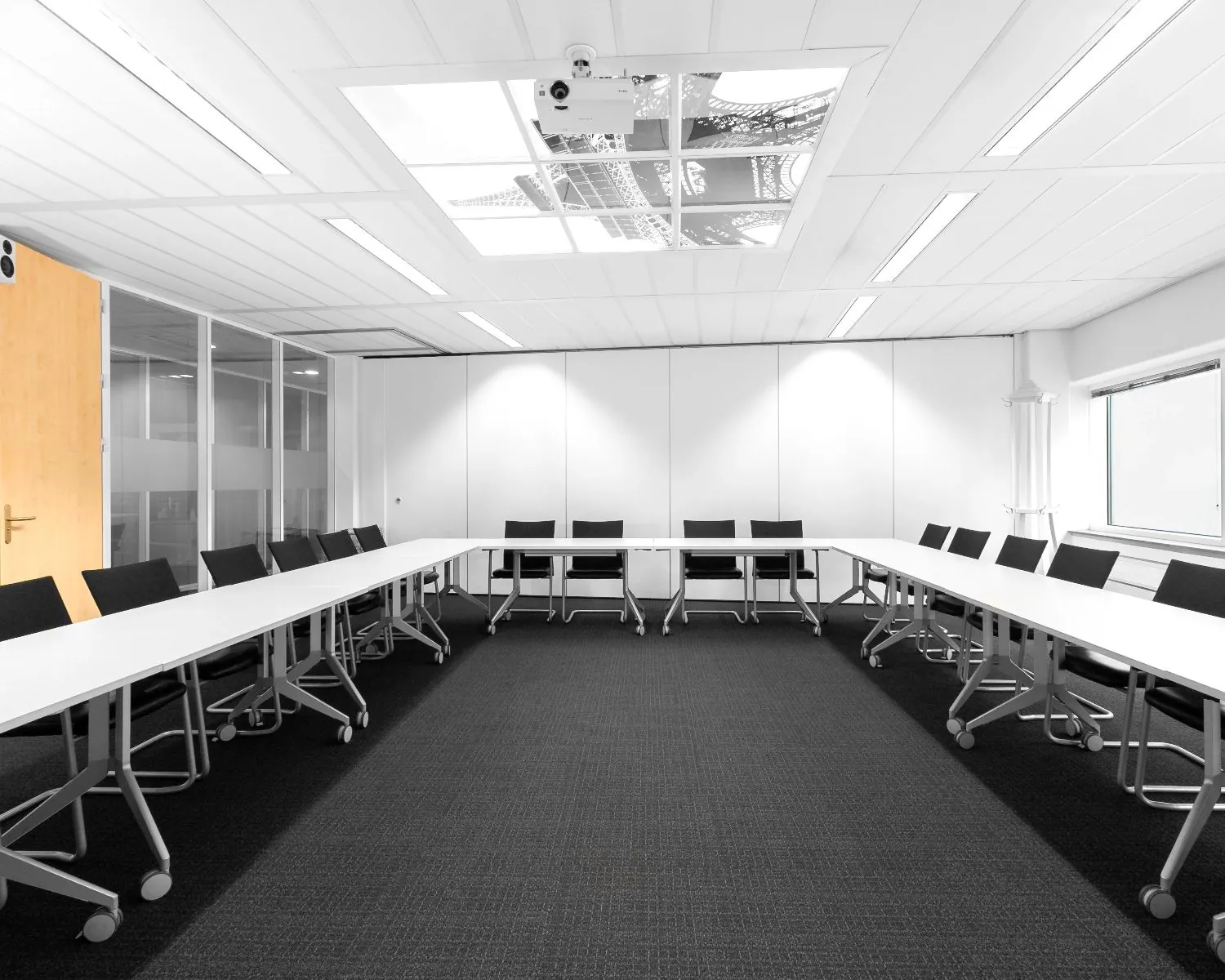 Moderne vergaderruimte met een U-opstelling van witte tafels en zwarte stoelen op wieltjes.